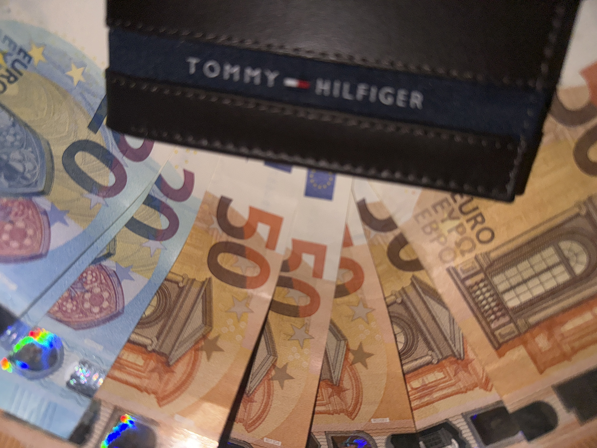 The image shows a black Tommy Hilfiger wallet on top of several euro banknotes. The denominations visible are 20 euro and 50 euro notes. The exact quantity of notes is not clear, as they are partially obscured by the wallet and arranged in a fan shape. The setting appears to be indoors, with a blurry background. The location (city/country) is not apparent from the image.
