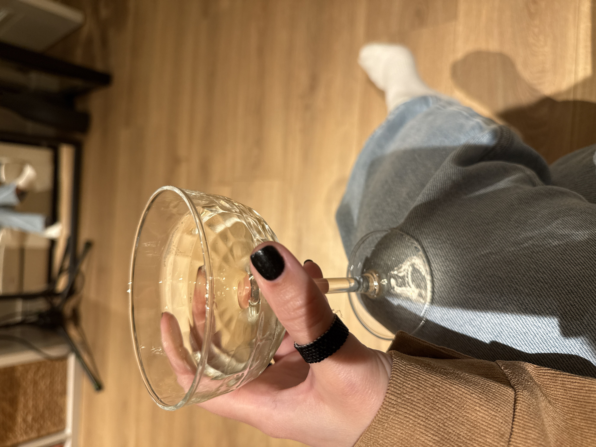 The image shows a person seated indoors, likely in a residential setting, with a partial view of their body. The person is wearing light-wash denim jeans, a brown corduroy shirt, and a white sock on their right foot. They are holding a coupe glass filled with a light-colored liquid. The person has black nail polish on their thumb, which is also adorned with a beaded ring.
The background consists of a wooden floor and a cabinet with some items on it. The lighting appears to be soft and diffused, possibly from natural light coming through a window. There is a shadow cast on the jeans and the floor. The location cannot be identified as Barcelona from the visual information provided.