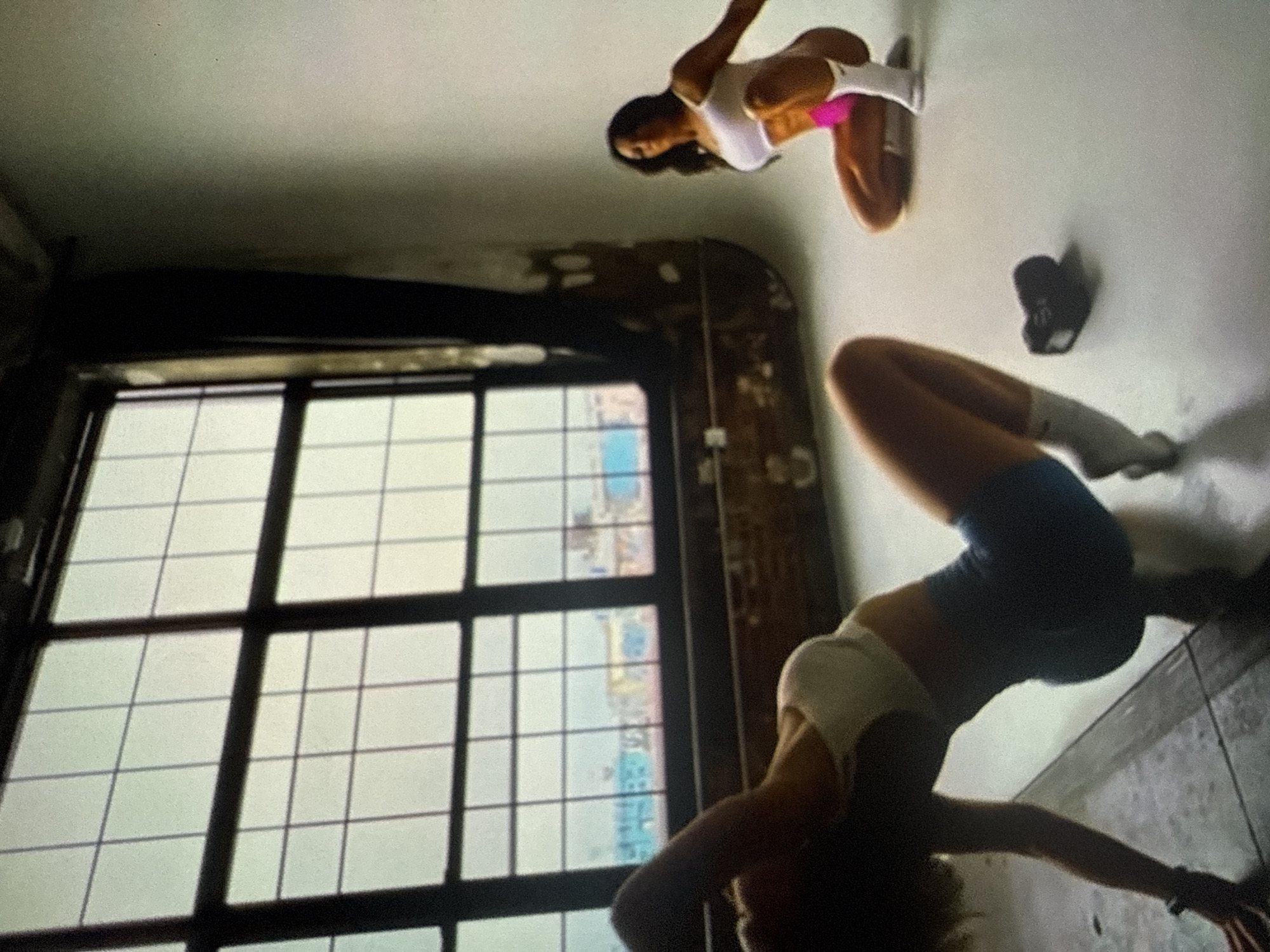 The scene is located in the city of Varna, Bulgaria, within what appears to be a studio space combining industrial architectural elements with a photography backdrop. Two women are present, both appearing to be in an athletic context.
In the foreground to the left, a woman with light skin and light hair is positioned on her hands and knees, arching her back. She wears a white sports bra, dark blue form-fitting shorts, and white athletic socks. A black wristband is visible on her left wrist. Her head is tilted downwards and to the left.
In the midground to the right, a second woman with darker skin and dark hair is kneeling on a white seamless floor. She wears a white sports bra, bright pink shorts, and white athletic socks. Her left arm rests on her left thigh, and her right arm extends slightly backward. She is facing towards the left of the frame.
Between the two women, a small black dumbbell rests on the white floor.
The environment features a large, multi-paned window occupying the left and upper-center portion of the background, revealing a distant urban landscape with discernible buildings painted in bright blue and yellow. Below the window, a section of exposed brick wall is visible. To the right of the window, the wall transitions into a plain, light-colored, seamless backdrop commonly used in photography studios. The floor under the first woman appears to be a darker, possibly concrete or tiled surface, which transitions to the white seamless floor under the second woman and the dumbbell.