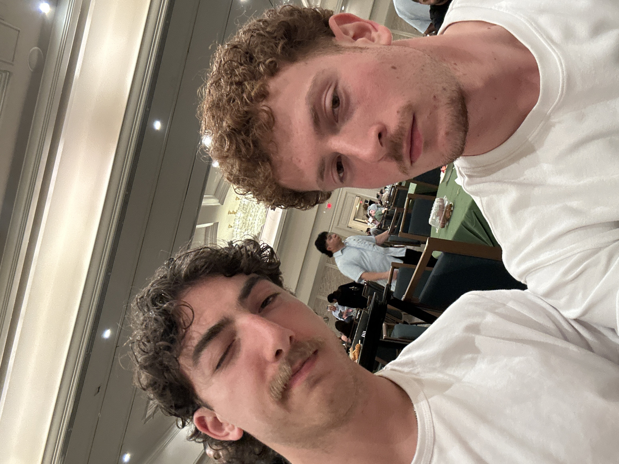 Two Caucasian males wearing white t-shirts are posing for a selfie. The male on the right has light brown, curly hair and a short beard. The male on the left has dark brown, curly hair with a mullet and a mustache. They appear to be in a large room with several rows of seating behind them. The room is well-lit with ornate light fixtures. In the background, other people are seated at tables. This scene appears to be inside the Aventura Turnberry Jewish Center, located in Aventura, Florida, United States of America.