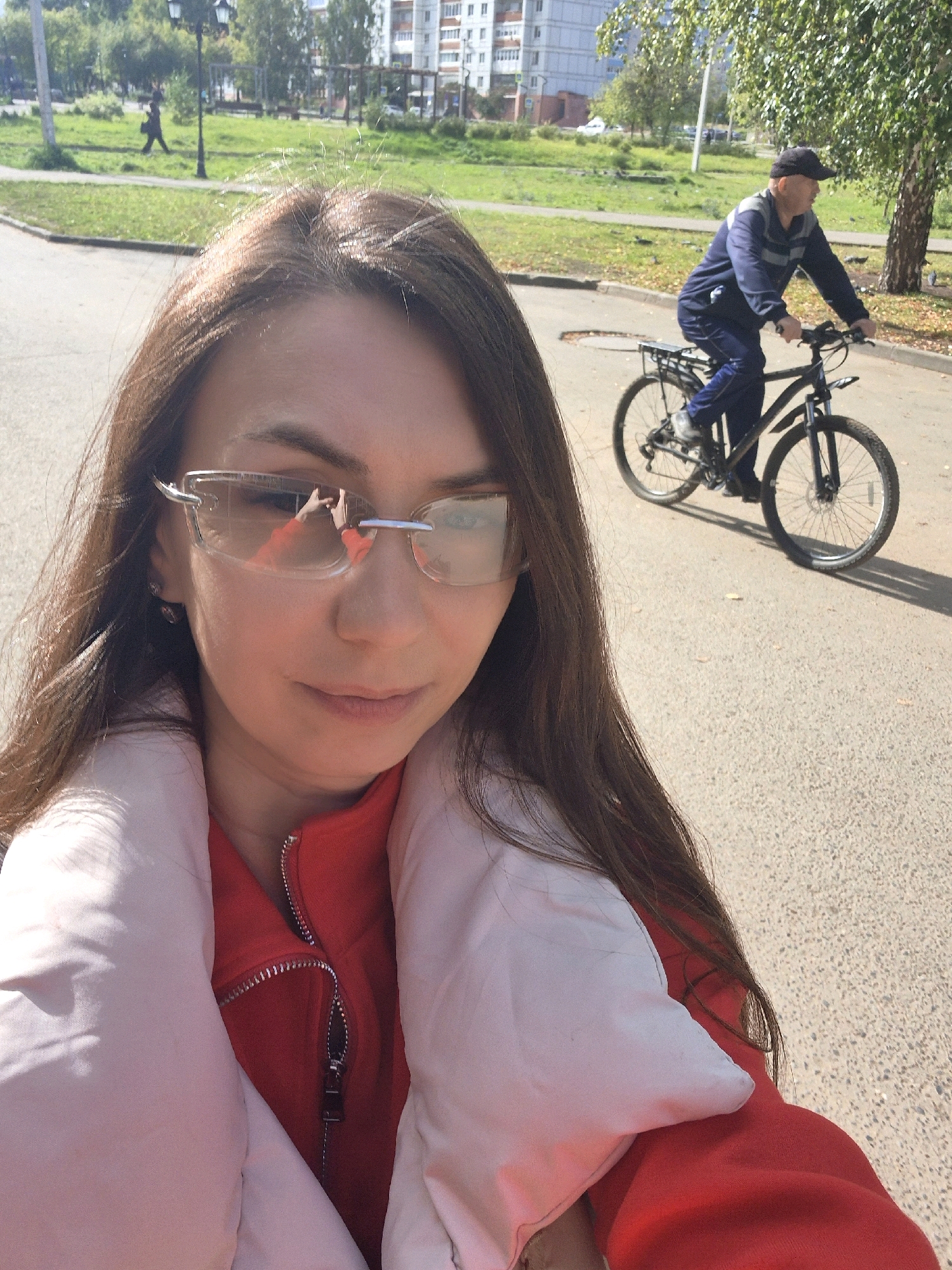 In Chaikovsky, Russia, a selfie is taken by a woman wearing glasses and a red jacket with a light pink puffer vest. She is in the foreground, with her face partially obscured by her hair and glasses. In the background, a man rides a bicycle on an asphalt road. The environment appears to be a park or residential area, with green grass, trees, and buildings visible. The lighting suggests it is daytime.