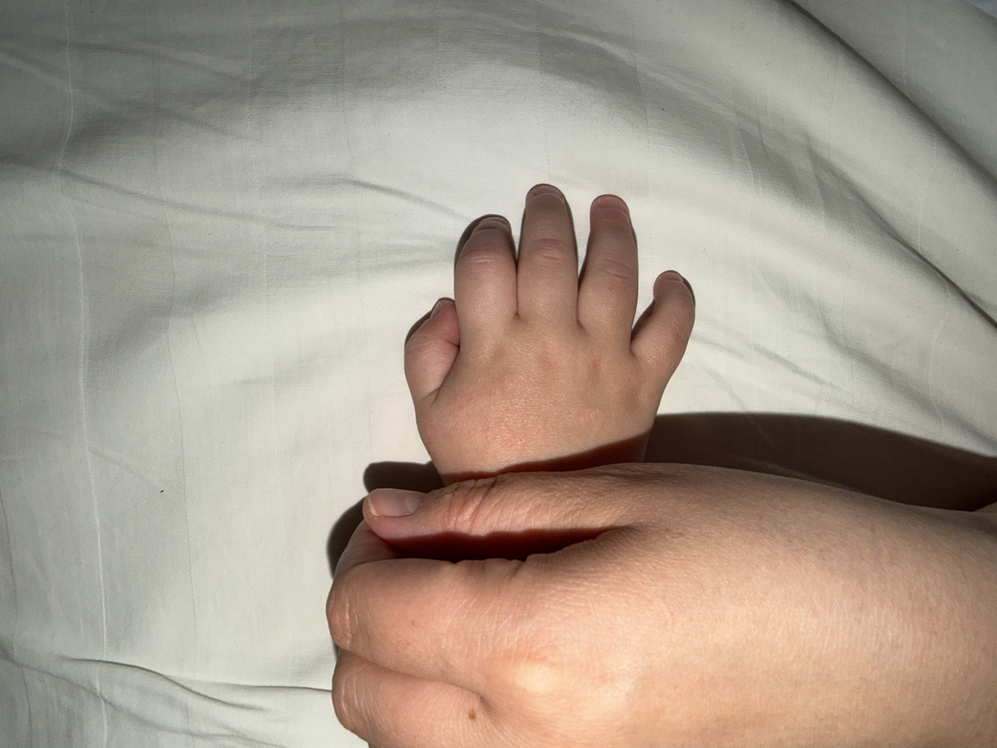 The image shows two hands on a white surface that appears to be a bed sheet. One hand is smaller, presumably a child's hand, with fingers slightly curled. The other hand is larger and appears to be an adult's hand, holding the child's wrist. No information in the image relates to the city of Chaikovsky, Russia.