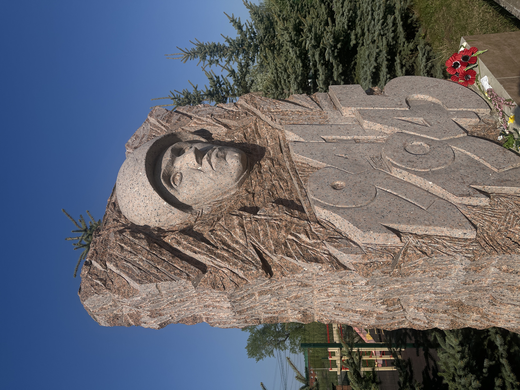 A monument is located in Фоки, Russia. The monument is made of stone. The face of a person wearing a helmet is carved into the stone. The year "1945" is carved into the stone as well. Some flowers have been placed on the monument. Trees are in the background. The sky is blue.