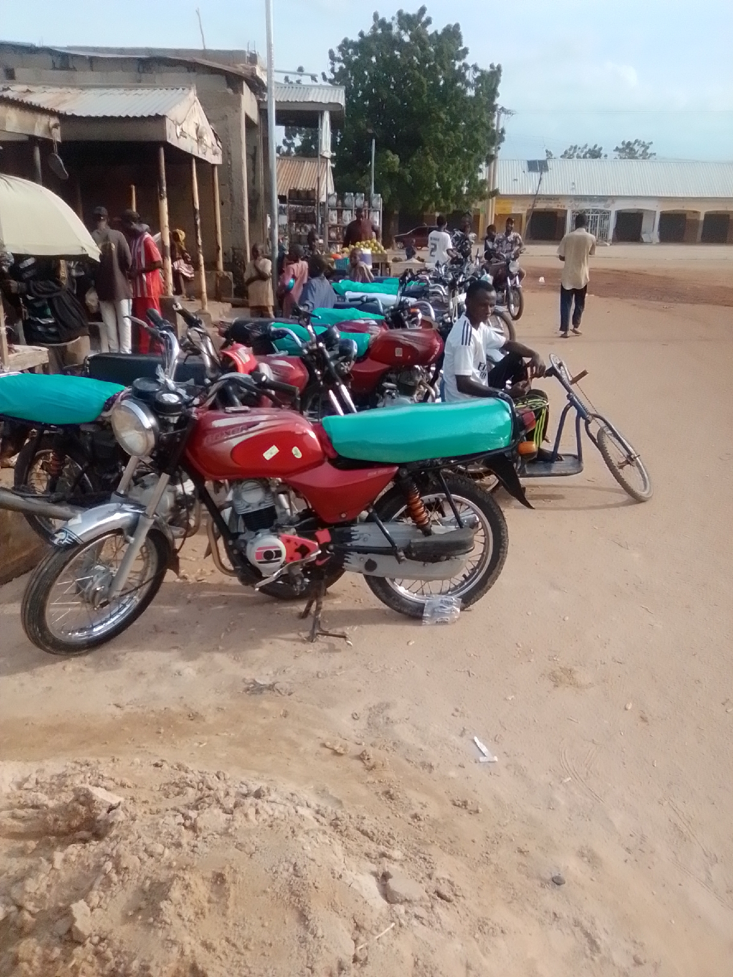 This is a medium-shot, eye-level, outdoor photograph taken in Galadima, Nigeria. The scene depicts a sandy marketplace with several motorcycles parked. In the foreground, a red motorcycle with a teal seat is prominently displayed. To its left, another motorcycle with a teal seat is partially visible. Behind these, a line of motorcycles extends further into the background. Several people are present, some standing and some seated on or near the motorcycles.
To the left of the frame, a section of a building with a corrugated metal roof and wooden support beams is visible, suggesting a market stall or shop. People are gathered under this structure. Further to the right, a long building with a white facade and arched openings lines the background. The ground is covered in reddish-brown sand, characteristic of the region. The sky is overcast with a pale, hazy light, indicating it might be early morning or late afternoon. A solitary tree with a full canopy stands in the mid-ground to the right. The overall impression is one of a busy, informal market setting in Nigeria.