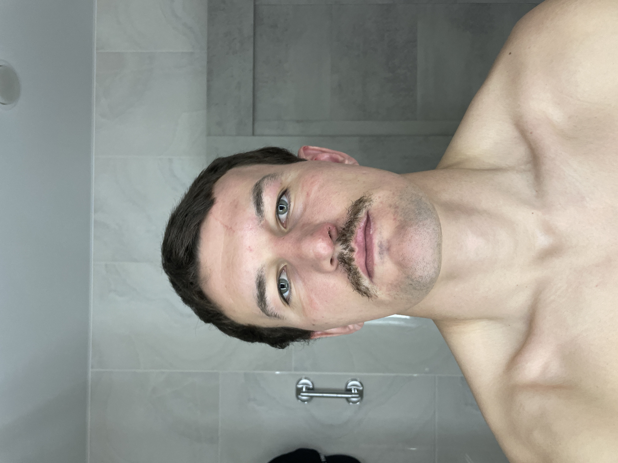 A medium, rotated, close-up shot depicts a shirtless man with a mustache, in what appears to be a bathroom. The background features tiled walls, a towel rack, and a partial view of what could be a dark garment on the floor. The location is not discernable from the visual information provided.