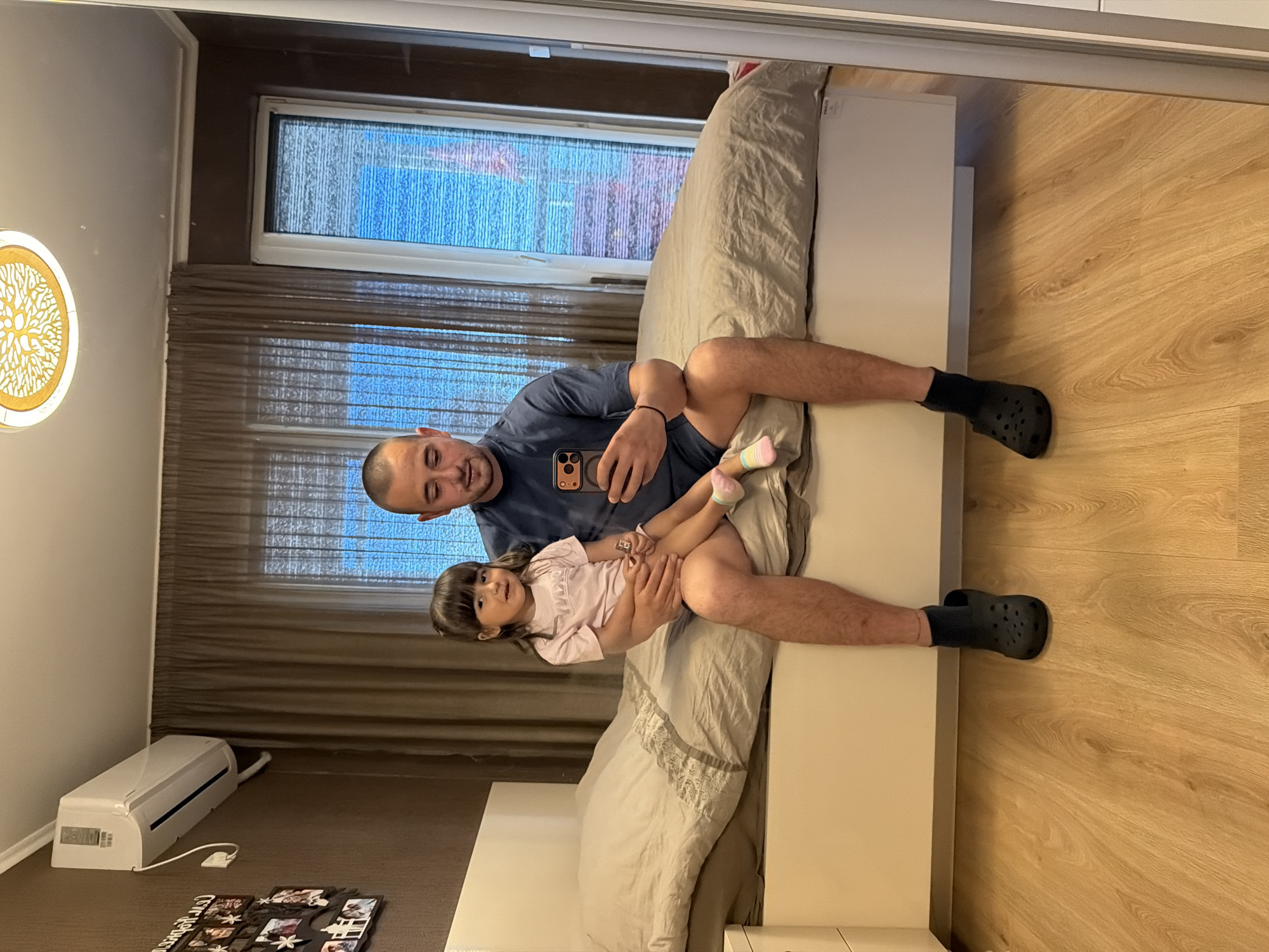 The image shows a man and a young girl sitting on a bed. The man is holding the girl on his lap and looking at a phone he is holding. The girl is looking towards the camera with a slight smile. They are both dressed casually. The man is wearing a dark blue t-shirt and black crocs. The girl is wearing a light pink t-shirt and colorful socks.
The scene appears to be indoors, likely a bedroom, with a bed, curtains, and a window visible in the background. A light fixture with a tree design is also visible on the left side of the frame, and an air conditioning unit is mounted on the wall below the window. The flooring is made of light-colored wood planks.
The overall mood of the image seems relaxed and intimate, suggesting a moment of connection between the father and daughter. The lighting suggests it might be daytime, possibly late afternoon or early evening, given the indoor setting and the light coming through the window. The photo was taken in Burgas, Bulgaria. There is no visible text in the image, apart from what might be on the air conditioning unit or the phone screen, which is not legible.