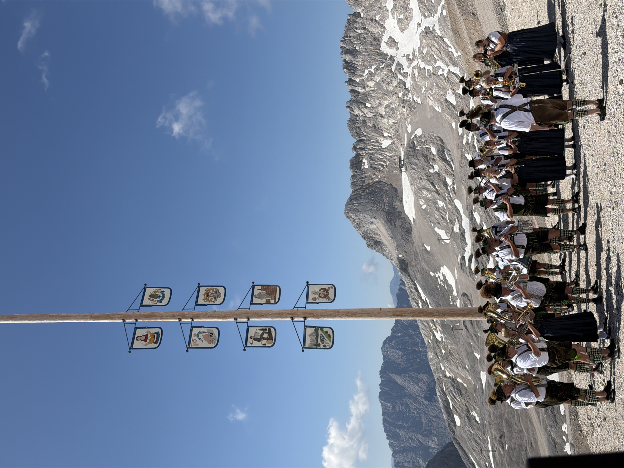 A band of musicians in traditional Bavarian attire plays brass instruments and clarinets on a rocky, mountainous terrain under a clear blue sky. The band members are adorned in lederhosen, suspenders, white shirts, and feathered hats. Behind them, a tall wooden pole is adorned with several rectangular banners, each featuring a painted image or crest. The background consists of jagged, snow-dusted mountain peaks. The scene takes place on a bright, sunny day. The location appears to be a high-altitude area, likely a mountain summit or a plateau, given the rocky landscape and sparse vegetation. While the image is set in a mountainous region, it is not explicitly stated to be in the city of Leutasch, Austria, though it could be a location associated with it.
