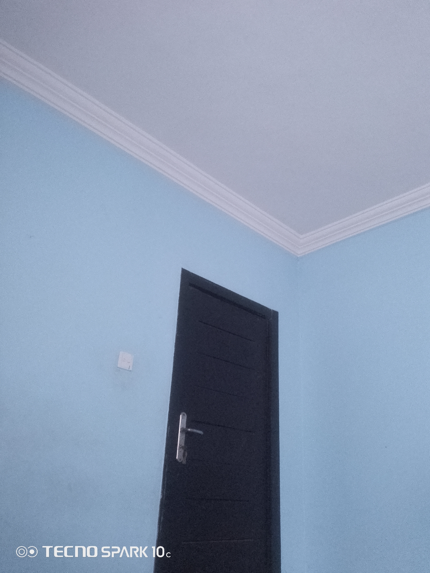 The image shows an interior view of a room with light blue walls and a white ceiling. A dark-colored door with a silver handle is visible on the right side of the frame. There is a small white electrical outlet on the wall to the left of the door. The TECNO SPARK 10c watermark is present in the bottom left corner. The scene depicts a simple, unfurnished room. The location is identified as Lekki, Nigeria.