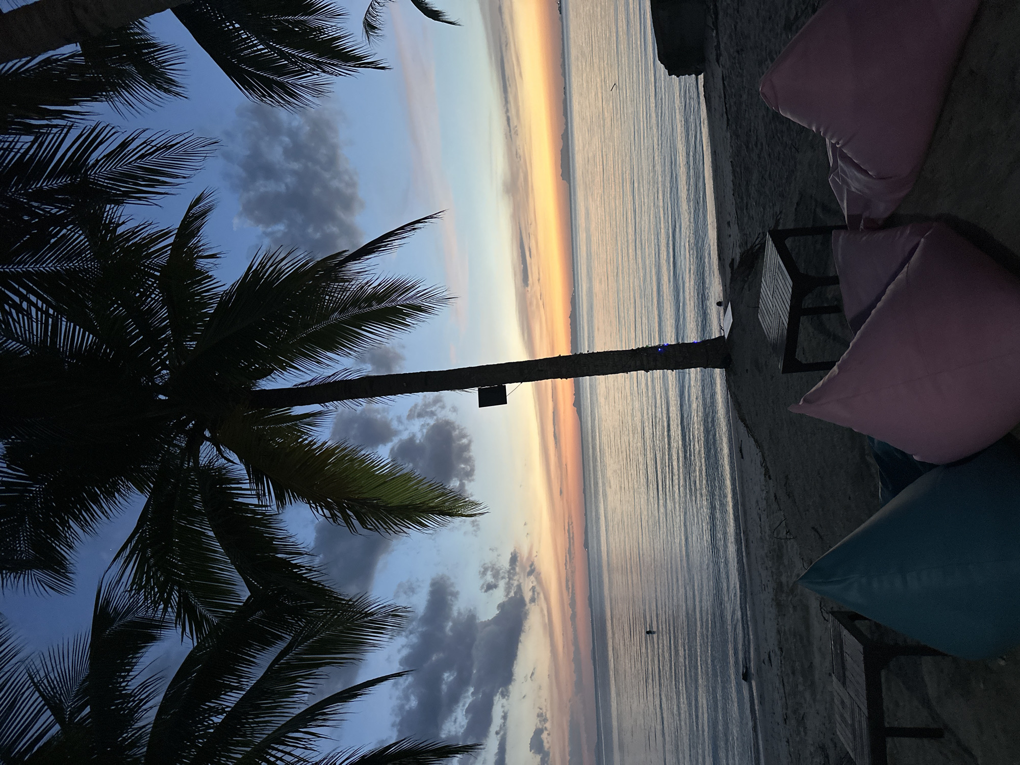 The image depicts a serene beach scene at dusk in Ko Samui District, Thailand. The sky is a gradient of blues and oranges, with scattered clouds reflecting the fading sunlight. The calm ocean stretches to the horizon, where distant islands are faintly visible. Palm trees frame the top of the image, their fronds silhouetted against the sky. In the foreground, beanbag chairs in teal and pink are set up on the sandy beach, along with a small wooden table, suggesting a relaxed seating area. The water's surface is gently rippled, and a single, barely discernible figure can be seen in the distance, wading in the sea. The overall atmosphere is tranquil and peaceful, characteristic of a tropical evening.