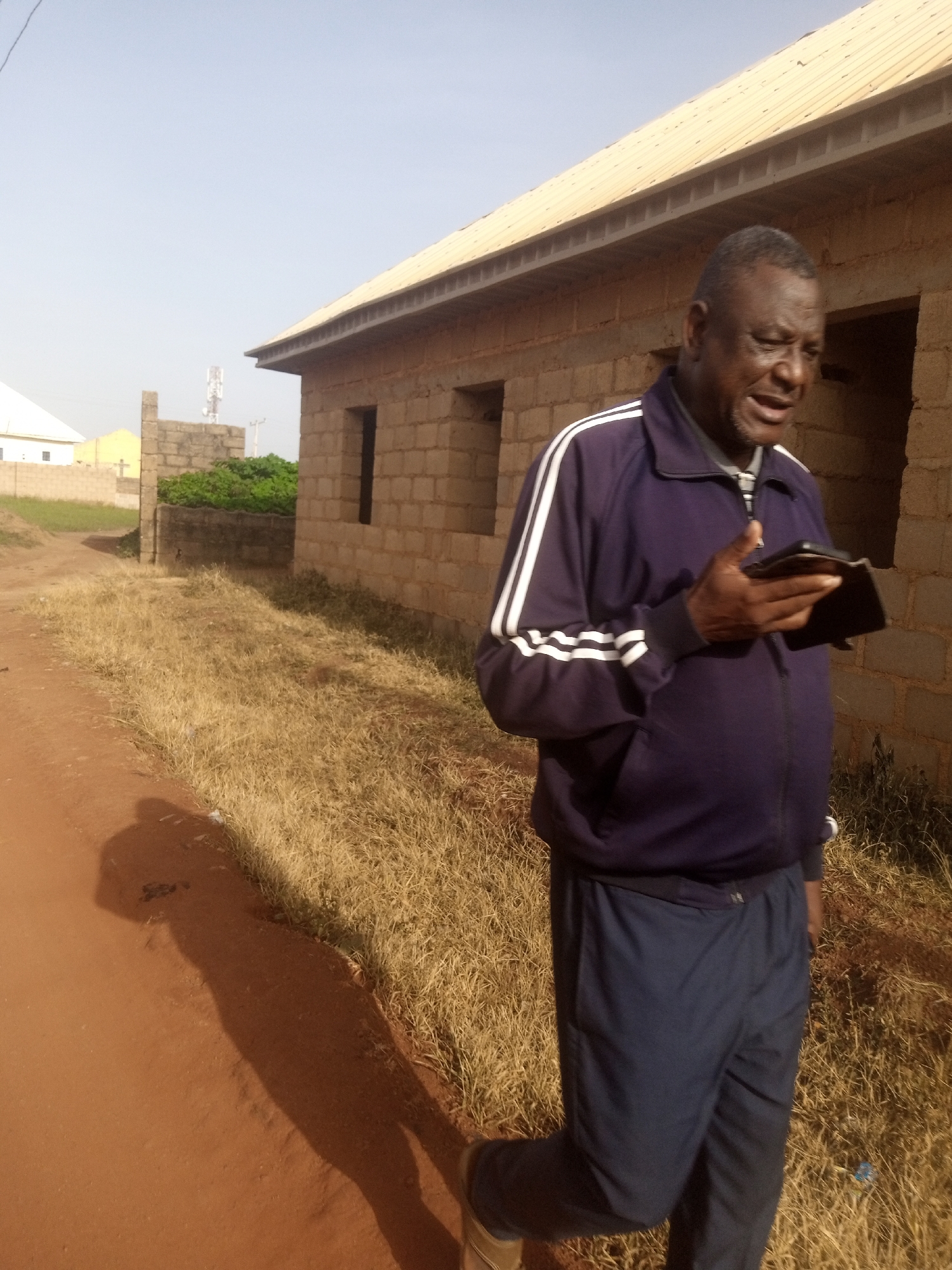 A middle-aged Black man, wearing a dark blue tracksuit with white stripes on the sleeves and dark blue pants, stands on a dirt path next to a field of dry grass. He is holding a dark rectangular object, possibly a tablet or e-reader, in his hands and appears to be looking at it. The man is in the foreground of the image. Behind him, under a light-colored metal roof, is a building constructed of light brown bricks with open doorways and window frames. The sky is clear and pale blue. In the background, a communication tower is visible, along with parts of other buildings. The scene suggests a semi-urban or developing area in Katon Rikkos, Nigeria.
