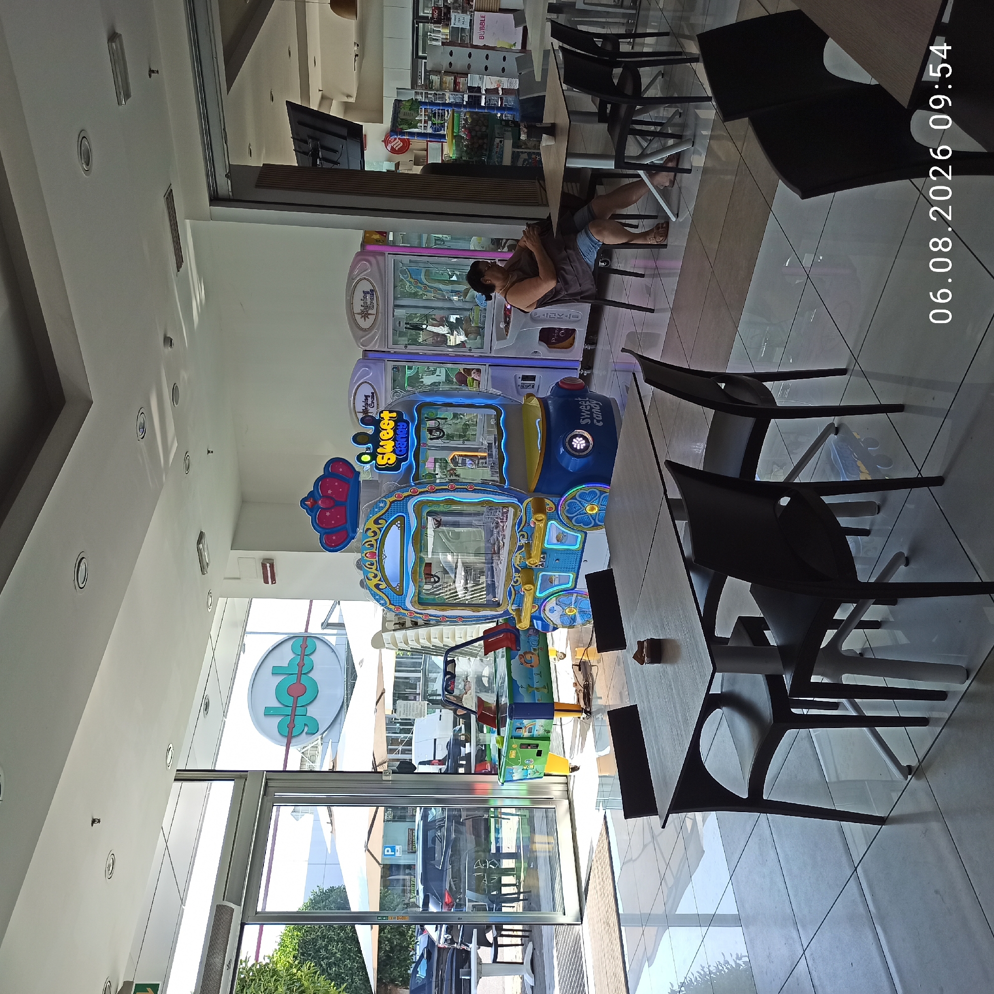The image depicts an interior scene of a retail establishment, likely a shopping center or a mall, with an outdoor view through large glass doors. Dominating the foreground are several tables and chairs, suggesting a seating or dining area. In the background, several arcade machines are visible, including a prominent "Sweet Candy" claw machine and other similar amusement devices. To the left, a sign with the word "globe" in a stylized font is displayed on a white oval background. Beyond the glass doors, an outdoor area with umbrellas and parked vehicles is visible, indicating a sunny day. A woman is seated at one of the tables, looking at her phone. The floor is tiled with a reflective surface. The timestamp "06.08.2026 09:54" is overlaid on the bottom right of the image.