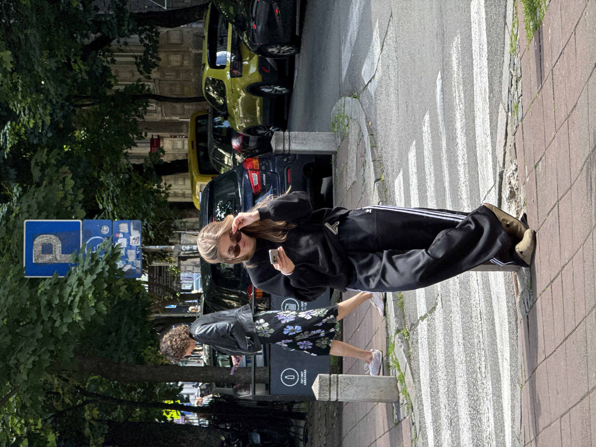 The image captures a street scene in Belgrade, Serbia, showing two women crossing a zebra crossing. One woman, wearing a black sweatshirt, tracksuit pants with white stripes, and tan-colored boots, holds her sunglasses on her forehead while looking at her smartphone. The other woman, walking behind her, is wearing a black leather jacket over a floral dress and pink sneakers. They are on a sidewalk made of pink bricks, next to a street with several parked cars. A parking sign is visible next to a tree on the left. A black advertising display is on the sidewalk.