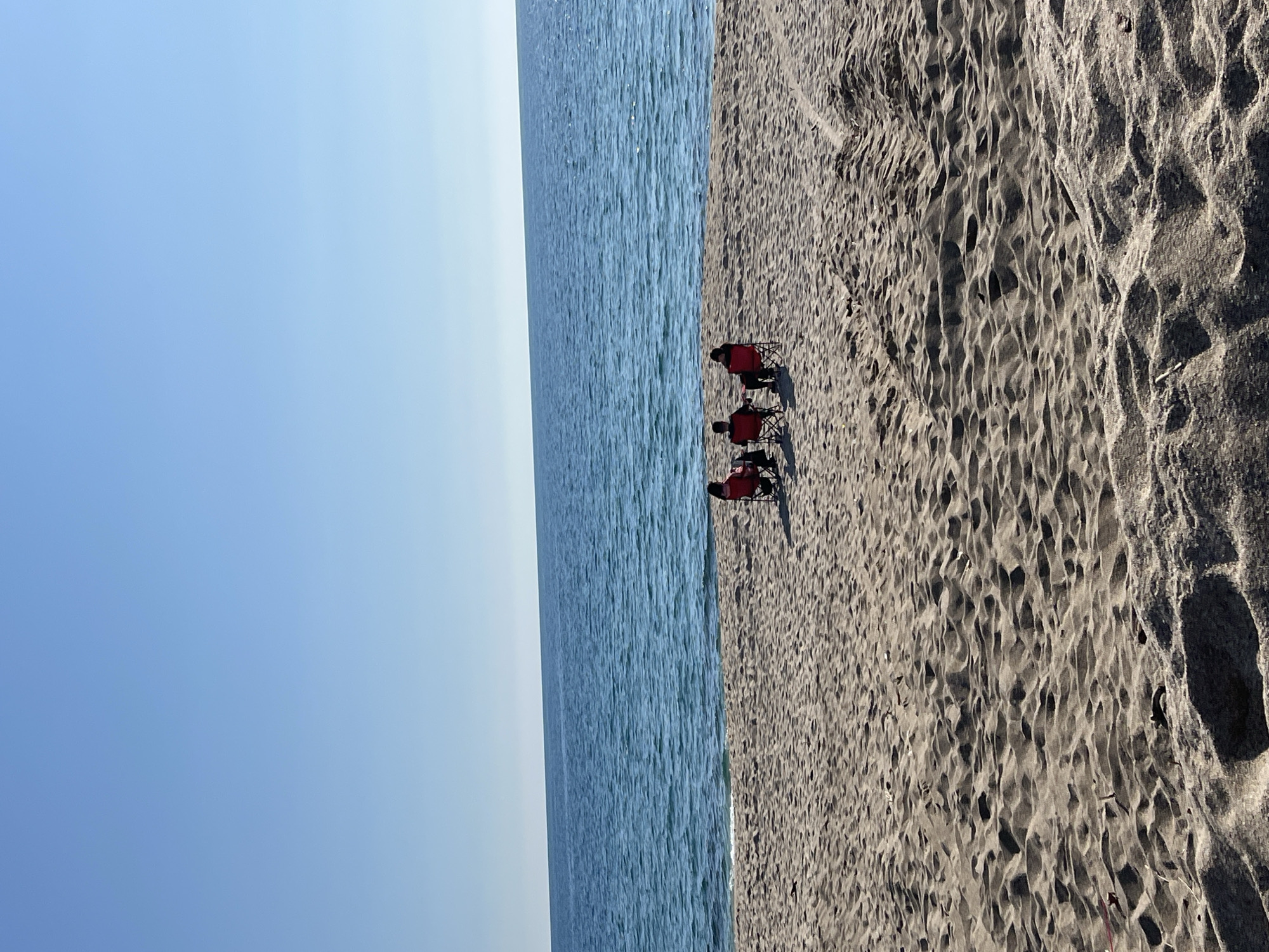 A vertical exterior shot captures three individuals seated in red folding chairs on a sandy beach in Alanya, Turkey, facing the Mediterranean Sea. The foreground consists of light-brown sand exhibiting pronounced ripple patterns, numerous footprints, and distinct tire tracks extending from the lower right. Small, dark specks are visible on the sand surface. The three individuals are positioned approximately in the middle ground, with their backs to the camera, looking out towards the water. The sea is a clear blue, displaying subtle wave action near the shoreline and extending to a distant, flat horizon line. Above the horizon, the sky is uniformly clear and light blue, devoid of clouds. The scene is illuminated by direct sunlight, casting shadows on the sand.