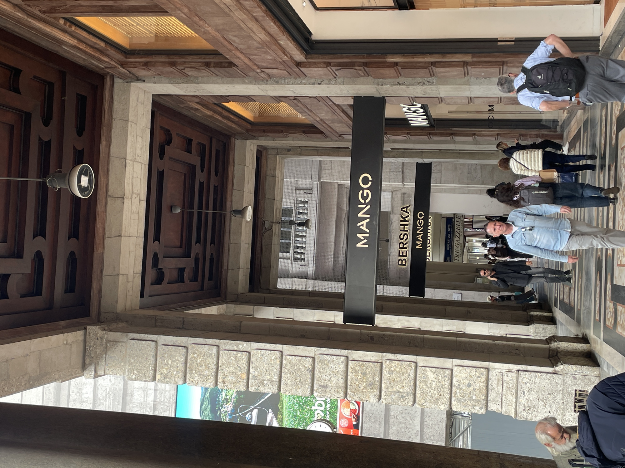 This image shows an indoor shopping arcade with a series of retail storefronts in Milan, Italy. The prominent stores are Mango and Bershka, indicated by hanging signs. The arcade features a covered walkway with tiled floors and stone pillars supporting the structure. The architecture includes ornate wooden detailing above the storefronts. Multiple people are walking through the arcade, some seemingly shopping and browsing. Daylight illuminates the space, and some reflective surfaces create highlights. The overall atmosphere is that of a bustling urban shopping area within a structured, architectural setting.