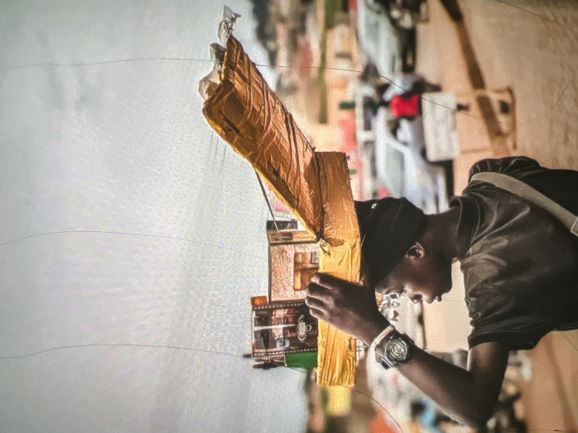 The image captures a street scene in Lagos, Nigeria, with a young person carrying a load of items balanced on their head. The individual, seen in profile from the shoulders up, wears a dark t-shirt and a black cap. A silver watch with a dark band is visible on their left wrist. The load on their head appears to be a collection of small boxes, possibly merchandise, partially covered by a yellow fabric and a large, crinkled golden sheet. The background suggests a busy urban environment with blurred out vehicles, buildings, and other individuals. The ground is a dusty orange-brown, indicative of a typical Lagos street. The sky is overcast with some lighter areas. The overall impression is of daily life and commerce in a bustling city.