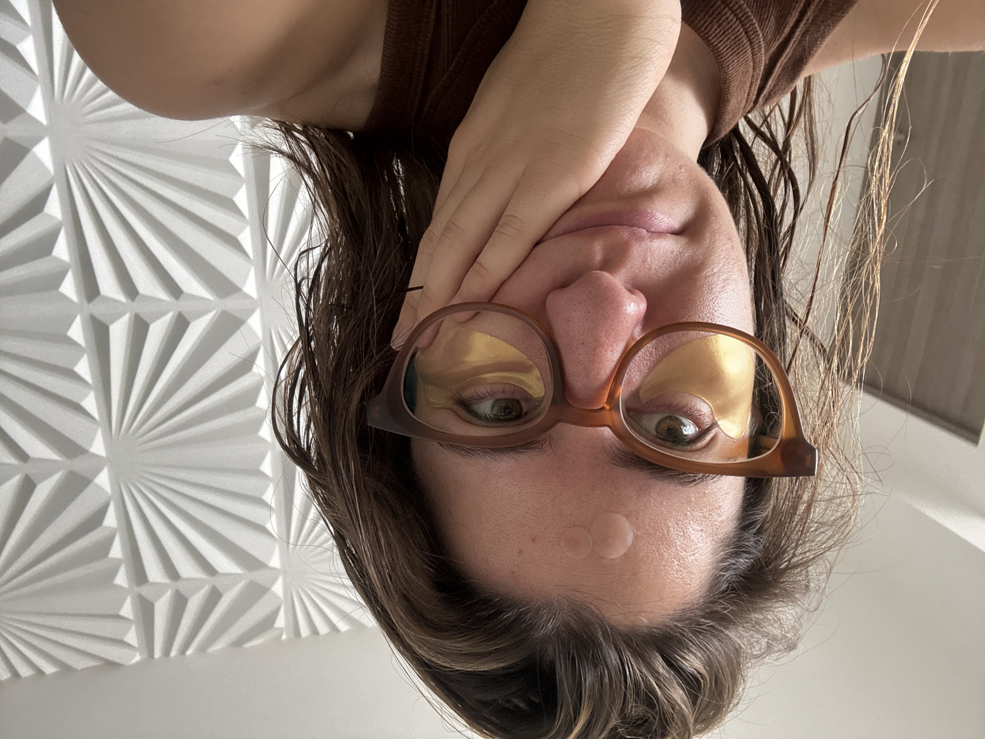 A selfie-style image from an unusual perspective shows a fair-skinned person with brown hair wearing eyeglasses. The person is holding their hand up to their face. There are two clear, circular patches on the forehead. Eye masks are visible under the eyeglasses. The background is a geometric wall with a fan pattern. The location is not identifiable as Puebla, Mexico, or any other specific location.