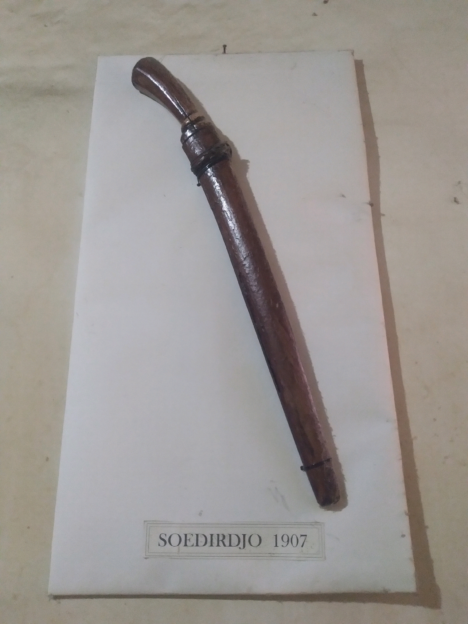 The image shows a wooden sheath with a curved top, possibly for a knife or sword. The sheath is dark brown and has a metallic band around its upper part, and another band near the bottom. It rests on a white surface, which is placed on a light-colored textured wall. At the bottom of the white surface, a label reads "SOEDIRDJO 1907" in dark text, enclosed in a thin border. The lighting is dim, casting shadows. The context suggests this is an exhibit, possibly a historical artifact. There are no people visible in the image. The location, Dukuh Klopo, Indonesia, is not directly depicted but is provided as context.