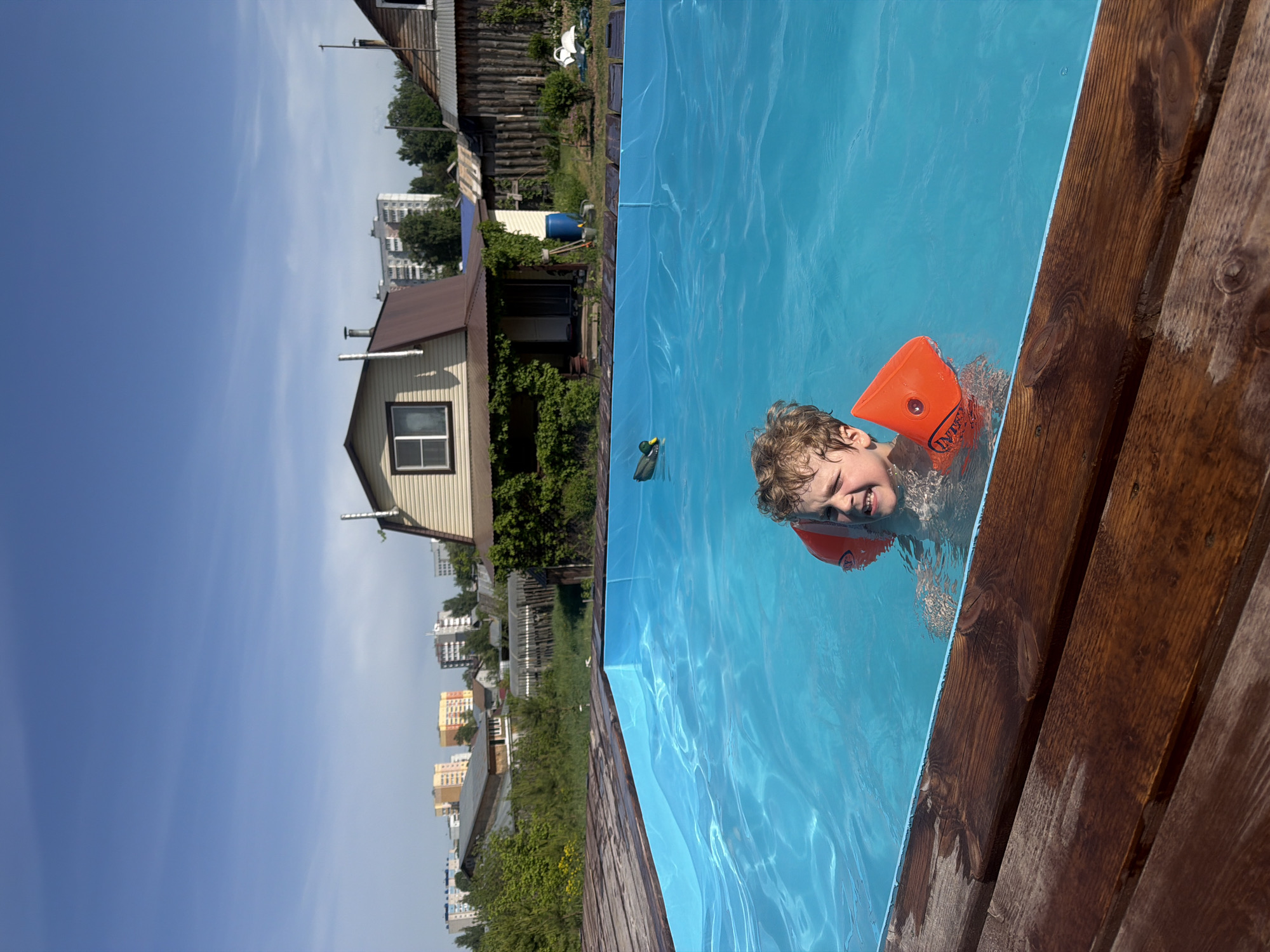 A boy is in a rectangular, blue, above-ground pool in Chaikovsky, Russia. The boy is wearing orange inflatable arm floats and is near the wooden edge of the pool. Behind the pool is a yellow, two-story house with a brown roof. Next to the house is a wooden fence and trees. In the distance are apartment buildings, and the sky is blue with some clouds.
