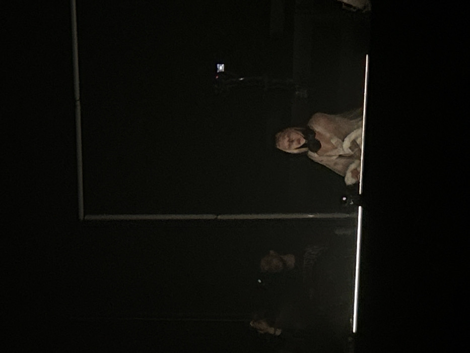 Two individuals appear in a dimly lit setting. One person, wearing headphones and wrapped in a light-colored garment, is positioned near a door or entrance with a bright vertical light strip. Below, another figure with a darker complexion seems to be bending down or kneeling. A rectangular frame or structure is partially visible, possibly indicating architectural elements within the space.