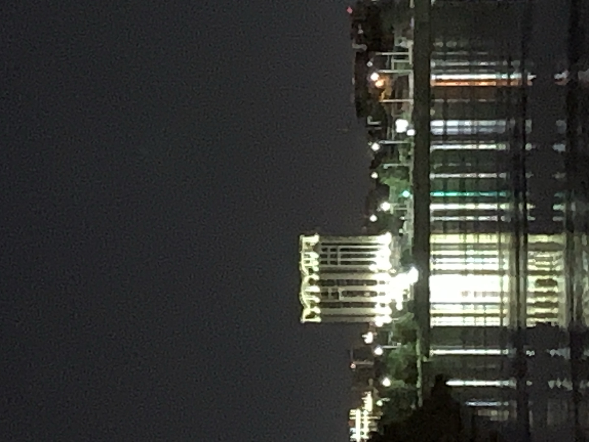 A nighttime, long-shot, exterior view of a city skyline shows a body of water in the foreground and a building with a distinctive arched roofline, illuminated by artificial lights, in the mid-ground. The building is tall and rectangular, with a series of vertical columns that appear to be lit from within or by external lighting. The arched roofline is also brightly lit, creating a decorative effect. The water reflects the lights from the building and other distant lights, creating a shimmering effect on its surface. The sky above is dark and appears to be clear. Other buildings and trees are visible in the background, also illuminated by lights. The overall impression is of a well-lit urban landscape at night, viewed across a body of water.