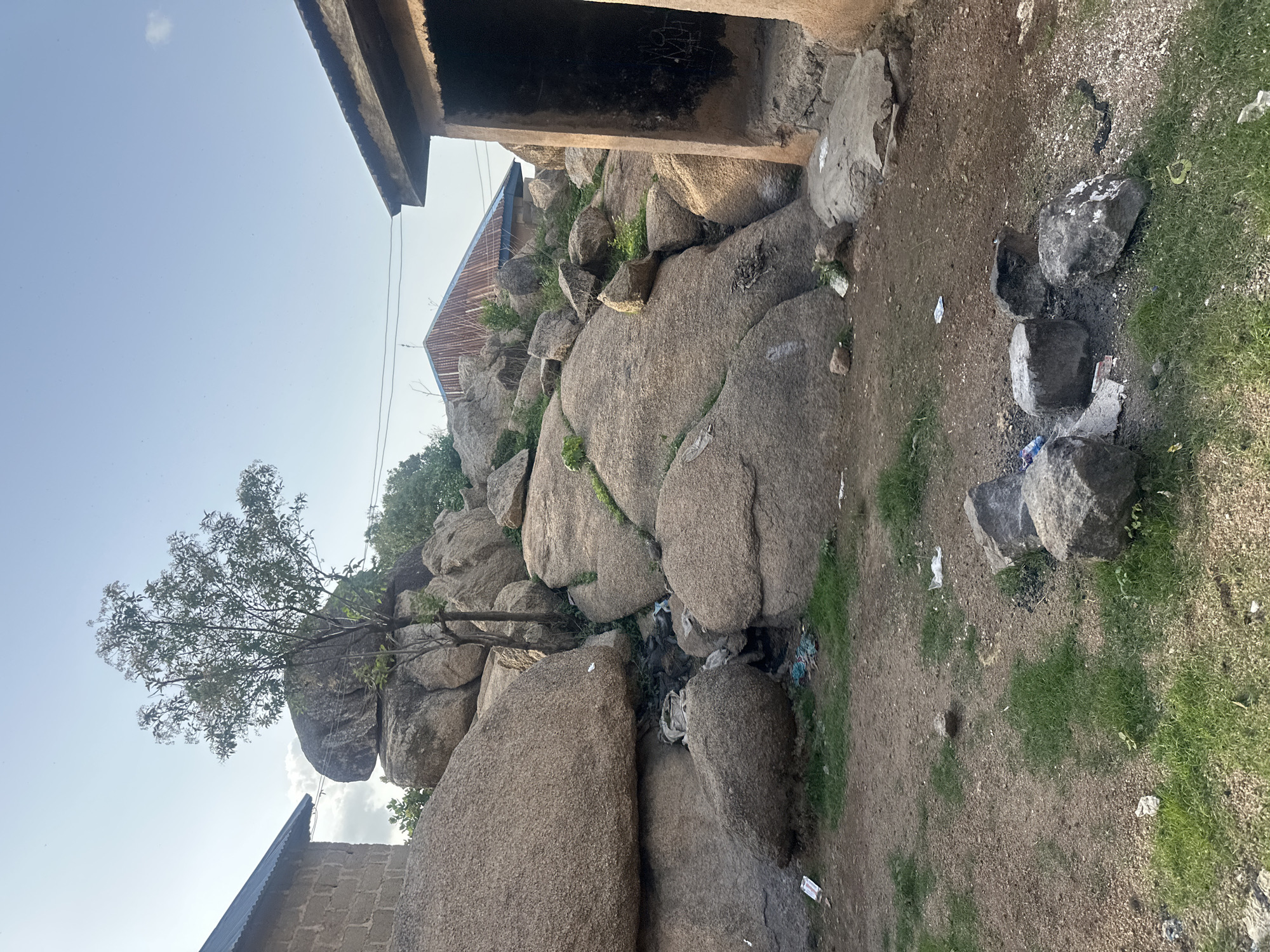 This image captures a scene in a residential area near a rocky outcrop in Jos, Nigeria. The foreground features dry, reddish-brown earth with patches of sparse green grass. Several large, grey rocks are scattered on the ground, with some appearing to be part of a makeshift fire pit, evidenced by ash and scattered debris.
In the mid-ground, a massive cluster of large, weathered granite rocks dominates the landscape, forming a natural embankment. These rocks are varied in size and shape, with some appearing rounded and smooth, while others are more angular. A young tree with sparse foliage grows precariously between the rocks. In the crevices and at the base of the rocks, there is visible accumulation of trash, including plastic bags and other discarded items.
To the left, a portion of a building constructed with light-colored bricks and a corrugated metal roof is visible, partially obscured by the rocks. To the right, another building with rough, earthen walls and a corrugated metal roof is prominent. The interior of this building appears open and dark, with some indistinct markings on the wall. In the background, more of the rocky outcrop and vegetation can be seen, as well as the roofs of other structures. The sky above is a pale blue with a few wispy white clouds. The overall impression is one of a somewhat untidy, rustic environment with significant geological features integrated into human habitation.