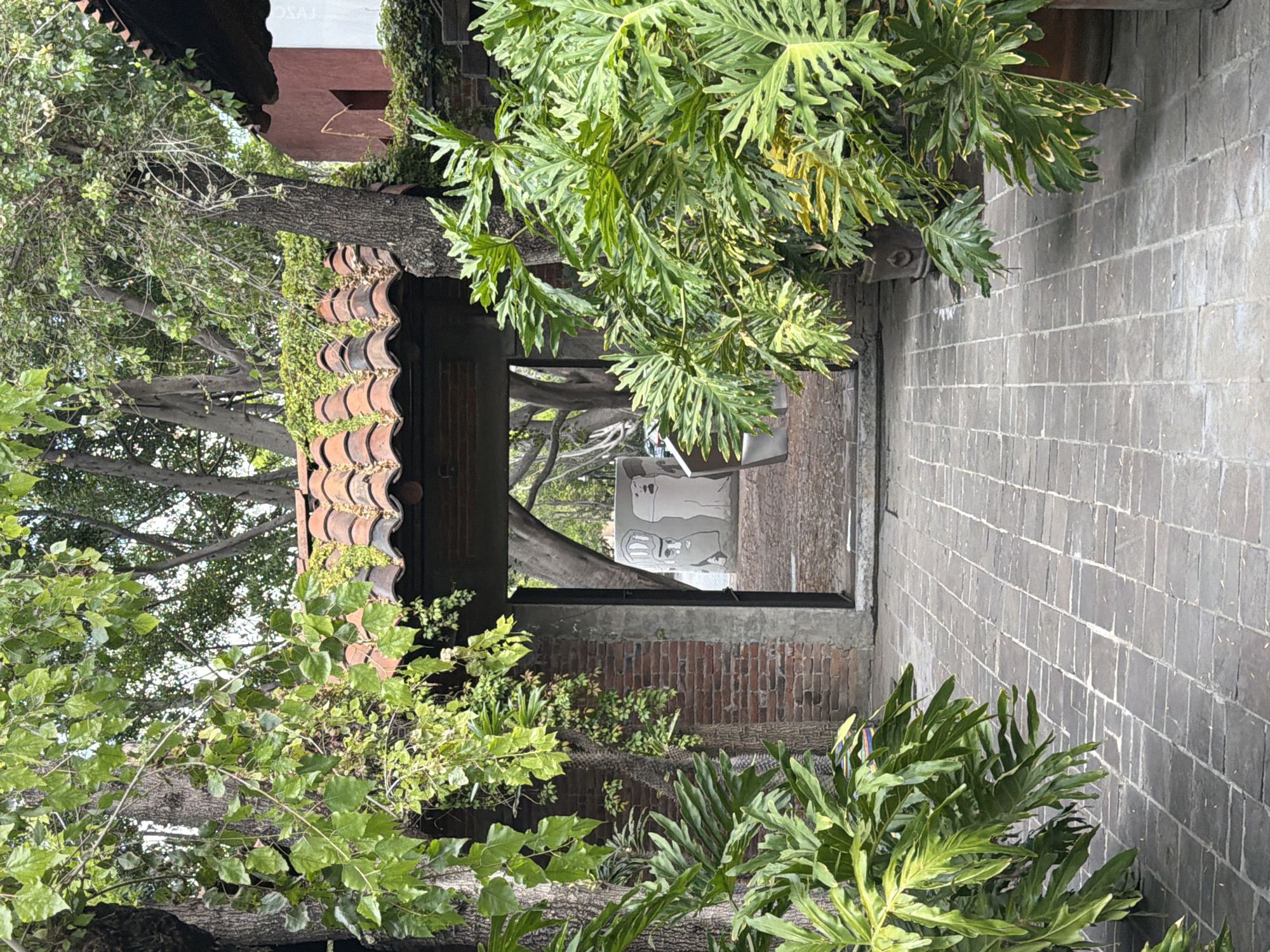 The image captures a perspective view through a covered passageway, flanked by lush greenery and a brick-paved path. The passageway is lined with brick columns and capped with terracotta tiles, creating a rustic, shaded area. Beyond the passageway, a glimpse of daylight and signage can be seen. The brick path is arranged in a linear pattern, leading towards the opening of the passageway. The scene suggests an outdoor setting in a place where brick and terracotta elements are common in architecture. This image is captured from Puebla, Mexico, in Puebla, Mexico.