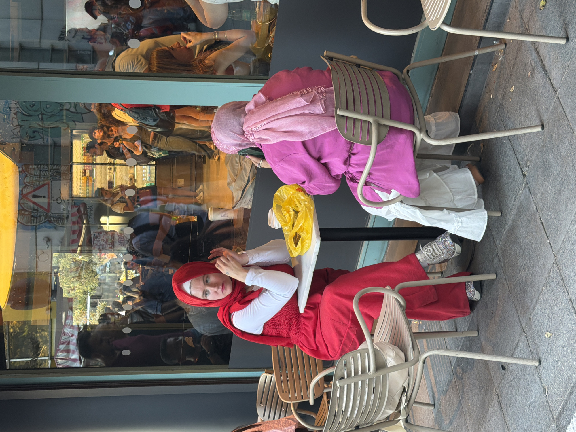 Two women, both wearing hijabs, are seated at a small square table outside a cafe in Kadıköy, Turkey. The woman on the left is wearing a red hijab and a matching red outfit, with a white long-sleeved shirt underneath. She is looking to her left with her hands clasped in front of her. The woman on the right is wearing a pink hijab and a pink tunic over a white ruffled skirt. She is looking down at the table. A bright yellow plastic bag is placed on the table between them. In the background, the reflection in the cafe window shows a busy street scene with pedestrians and shops. The pavement is made of grey cobblestones, and metal chairs are set around the table.