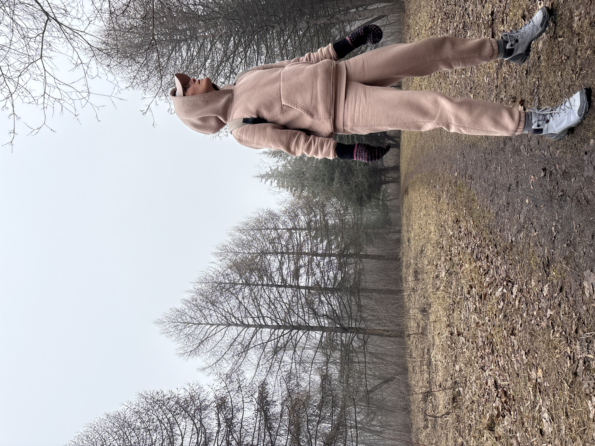 A person wearing a light pink tracksuit, grey sneakers, and dark colored gloves stands in an outdoor area that appears to be a park or wooded area in Ulyanovsk, Russia. The person is looking up toward a cloudy sky and trees with bare branches are visible. The ground is covered in fallen leaves and vegetation. The overall atmosphere is foggy and muted in color.