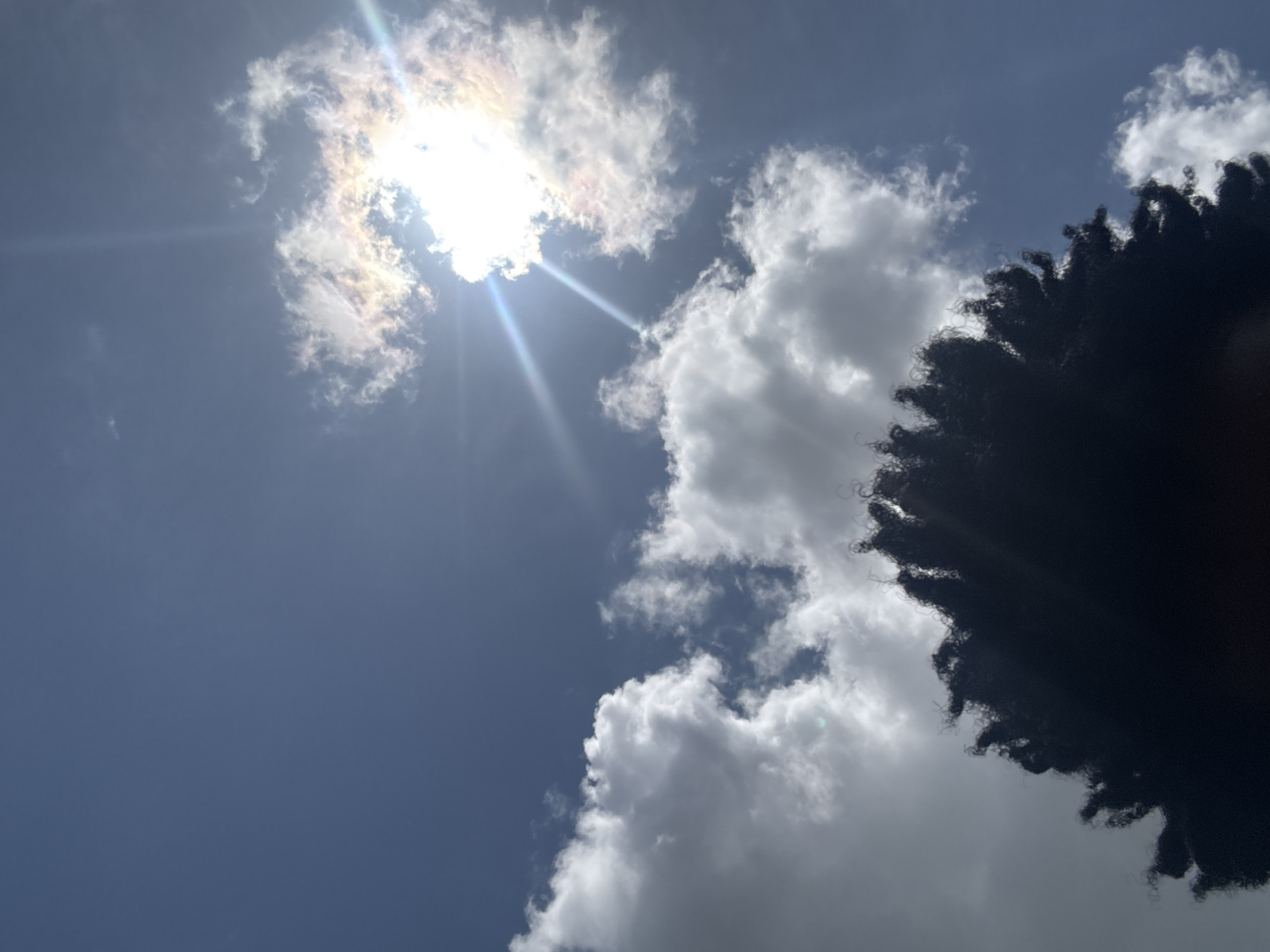The image shows a sky view taken in Ihiagwa, Nigeria. The sky is light blue with scattered white clouds. The sun is visible, partly obscured by a thin layer of cloud, with light rays emanating from it. A portion of a dark-colored, textured object, possibly hair, is present in the right side of the frame.