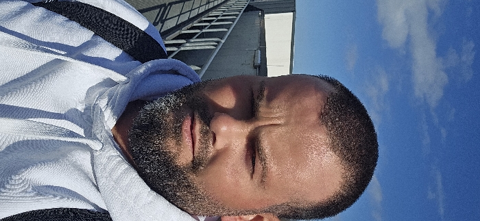 A vertical, eye-level photograph depicts a male subject outdoors in Delbrück, Germany, on a brightly lit day. The male, positioned centrally and filling most of the frame, looks directly at the camera with a neutral to slightly furrowed expression, suggesting possible squinting due to sunlight. He has short, dark, closely-cropped hair and a full beard with visible grey hairs. His skin appears tanned, with highlights on the forehead and cheeks indicating strong overhead or frontal illumination. He is wearing a white hooded sweatshirt with the hood down and visible drawstrings, and a black strap, likely from a backpack, is visible over his left shoulder.
In the upper left quadrant of the background, a metal railing, possibly part of an elevated walkway or bridge, is visible, extending horizontally. Beyond the railing, a light-colored industrial or utility building with a flat roof and white facade sections can be seen. The sky above is clear blue with sparse white cumulus clouds. The strong sunlight casts distinct shadows on the subject's face, particularly under his brow and nose.
