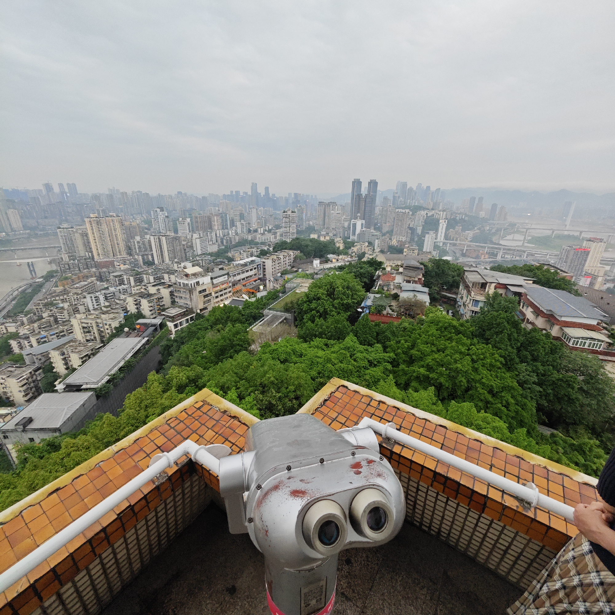 The image shows a cityscape in Yuzhong District, China, taken from an elevated vantage point. In the foreground is a coin-operated binocular viewer mounted on a tiled base. The binocular is silver and shows signs of wear. To the right edge of the frame, there is a glimpse of a person's hand and clothing; the person is partially out of frame.
Behind the binocular is a dense urban landscape featuring a mixture of high-rise buildings, residential structures, and green spaces. The architecture is varied, with both older and modern buildings visible. Trees and other vegetation are clustered in several areas, softening the urban sprawl. A large river and multiple bridges can be seen weaving through the city. The sky is overcast, lending a gray tone to the overall scene and slightly obscuring the distant buildings.