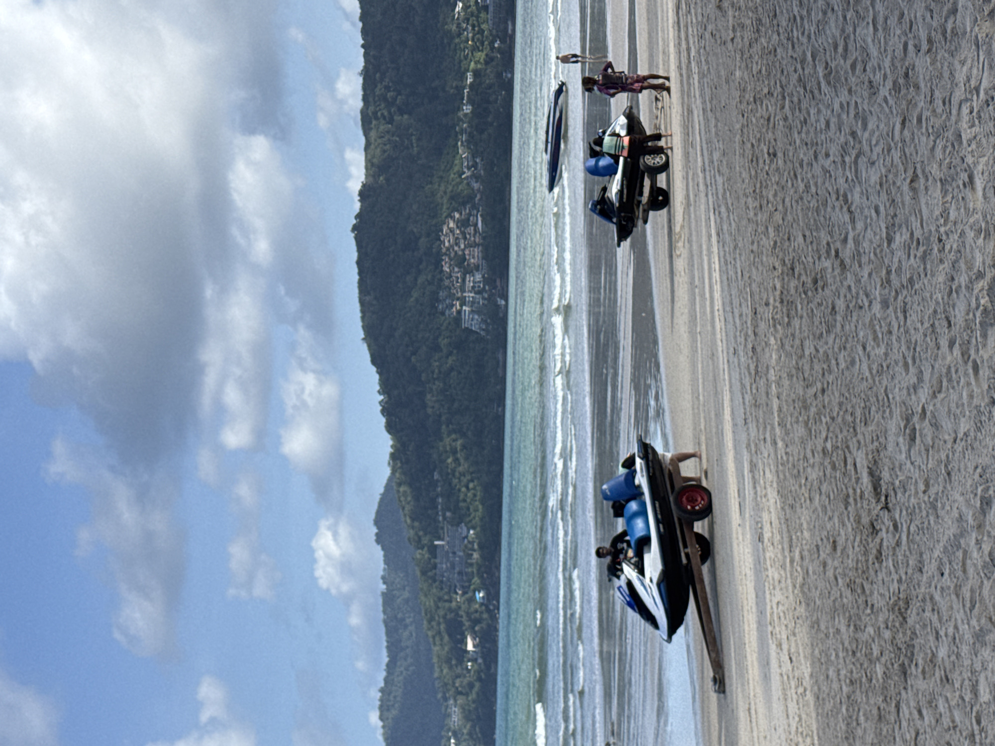 Two jet skis are on the beach in Patong, Thailand, one on a trailer with a person seated on it and another on the sand with two people standing nearby. In the background, the ocean waves are visible with a boat in the water, and lush green hills with buildings are on the horizon under a cloudy blue sky.