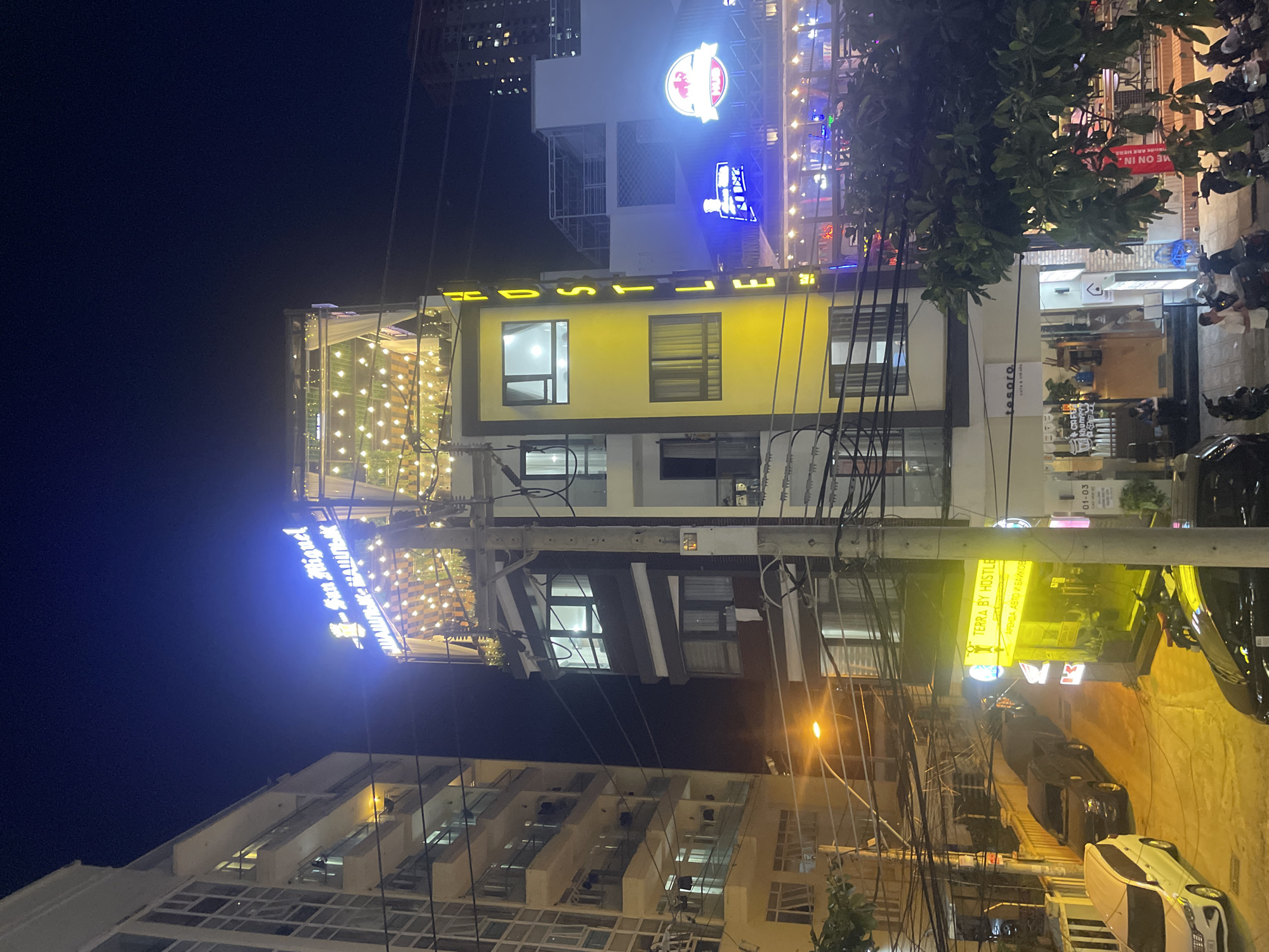 This night-time image captures a lively urban street scene in Bắc Nha Trang, Vietnam. The sky is dark, illuminated by numerous artificial light sources from buildings, signs, and streetlights.
A prominent multi-story building, likely a hostel, dominates the center. Its facade features a bright yellow section with windows, and above it, a large blue neon sign spells out "HOSTEL". Below, another sign reads "TERRA BY HOSTLE" and "АРЕНДА АВТО И БАЙКОВ" (Russian for "Car and Bike Rental"), indicating accommodation that also offers vehicle services. The building's upper levels are adorned with extensive string lights, creating a festive ambiance, possibly on a rooftop or terrace.
Various other businesses are identifiable by their illuminated signs. To the left, a "San Miguel" sign suggests a bar or restaurant. Further up, a circular red and white logo is visible. On the right, partially obscured by a tree, a sign reads "COME ON IN." and "We ARE HERE". Below, a smaller sign for "tesoro. CAFE" can be seen. Another sign further down the street advertises "ШАШЛЫК НАШЛЫК" (Shashlyk Nashlyk), "CAFE," and a number "01-03."
The street is busy with parked cars and numerous motorbikes, reflecting typical Vietnamese city life. Overhead, a complex network of power lines connects to a utility pole. The scene depicts a bustling commercial area, active into the night, with a mix of hospitality and food establishments. No specific individuals are clearly visible in the frame.
