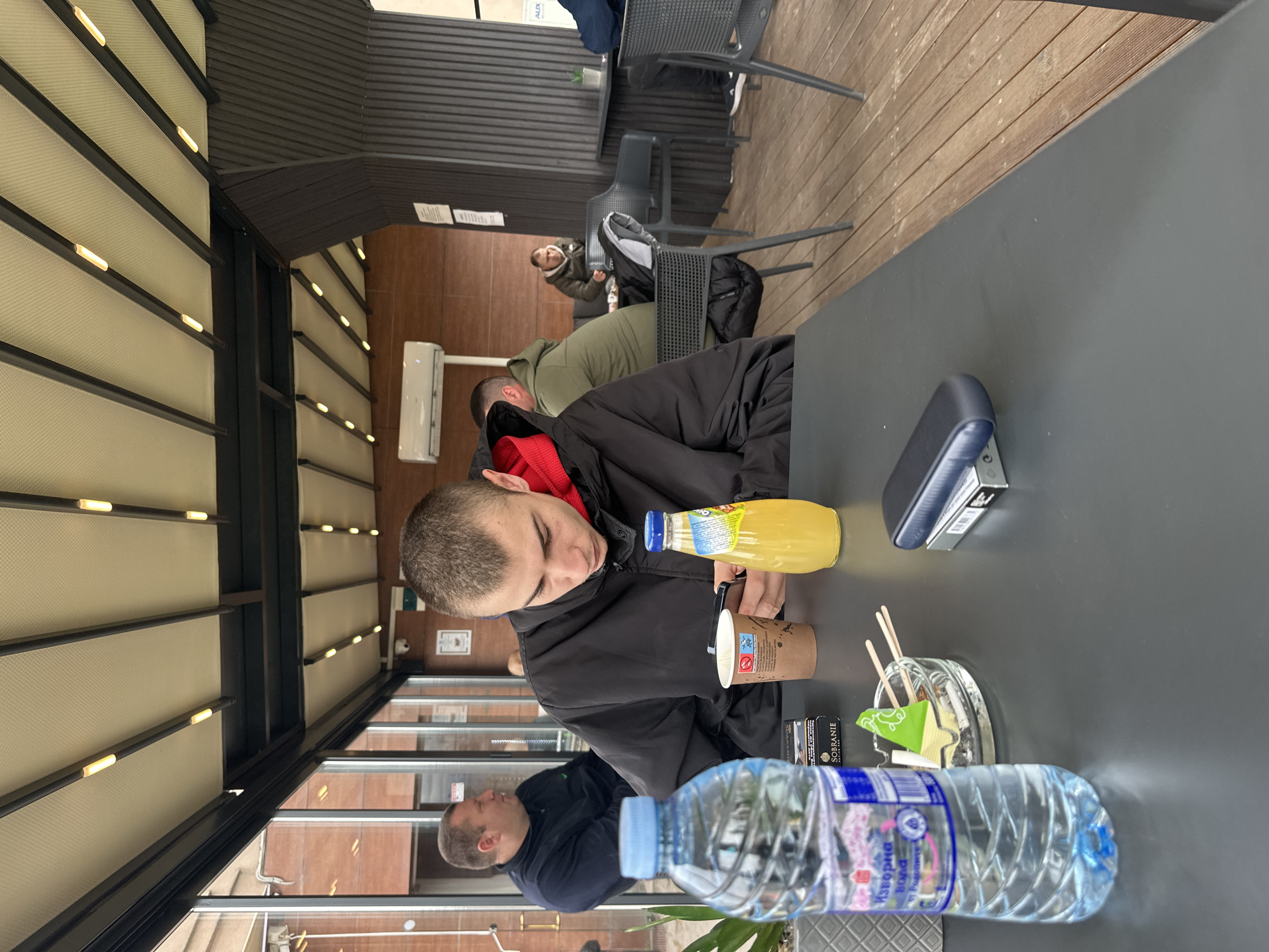 The image depicts a scene at a restaurant or cafe, possibly in an outdoor seating area, judging from the awnings and outdoor furniture. A young man wearing a black jacket and red hoodie is seated at a table. On the table are a bottle of water with Cyrillic lettering, suggesting the location is in Bulgaria, a bottle of juice, a cup (likely with coffee or tea), a stapler, an ashtray with cigarette filters and two packs of Sobranie cigarettes. A man in a green jacket is nearby. The architectural style and specific details suggest the location might be in Plovdiv, Bulgaria. Other people are visible in the background, seated and walking around. The general atmosphere suggests a casual dining or cafe setting.