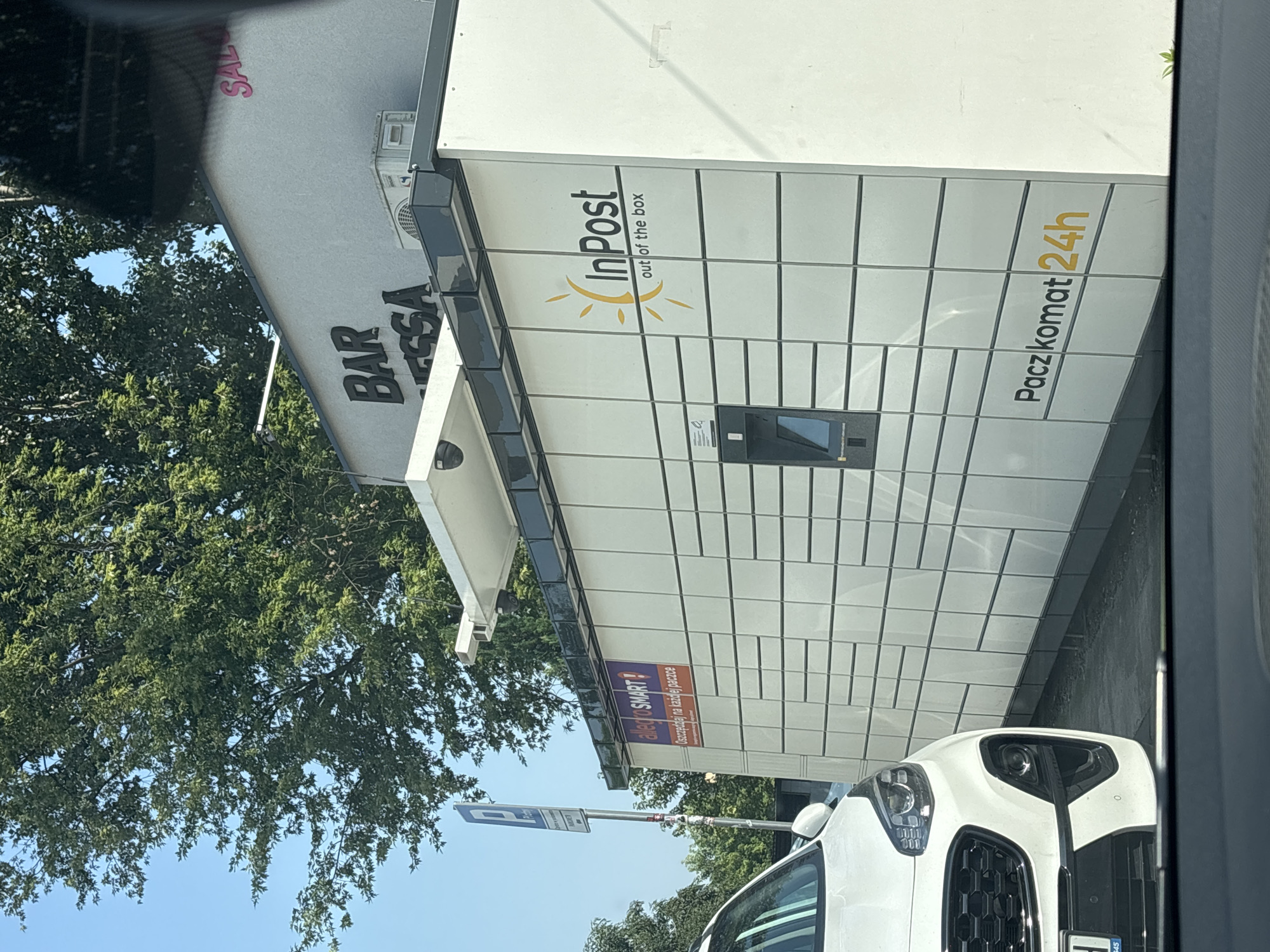 The image captures a street view in Krakow, Poland, focusing on an InPost parcel locker station. A white SUV is partially visible in the foreground, parked next to the parcel locker. The parcel locker is a modular, white structure with numerous individual compartments, some of which are open or closed. The InPost logo, featuring a stylized sun and the text "InPost out of the box," is prominently displayed on the side of the locker. Above the parcel locker, a building with signage for "BAR" and "SAL" can be seen, along with an outdoor air conditioning unit and security cameras. A blue parking sign is also visible on a pole to the left. The environment suggests an urban setting with trees and a clear sky.