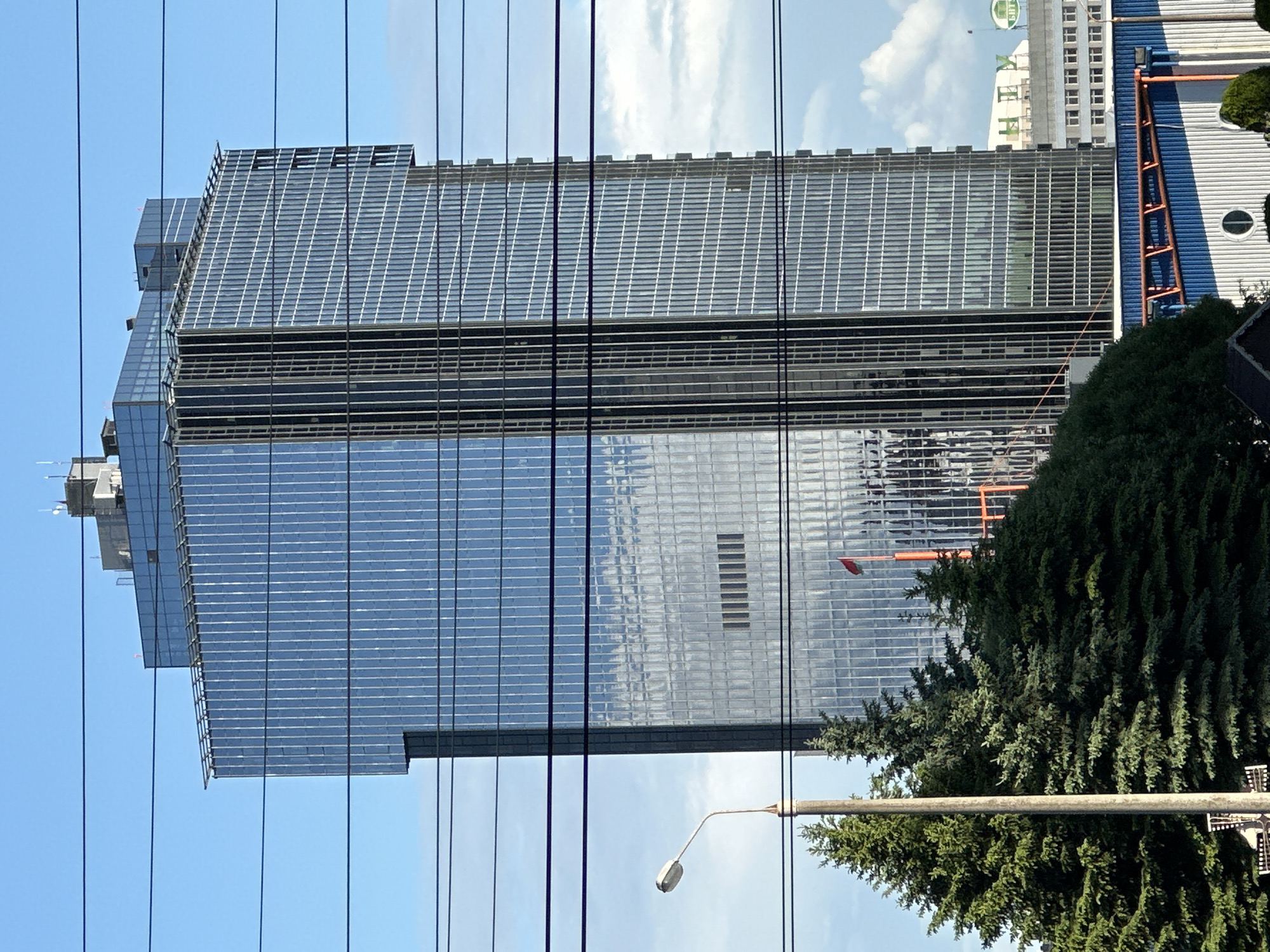 A tall, modern glass-clad building dominates the scene, likely located in Minsk, Belarus, given the context. The building features a grid-like façade and a stepped design at its upper levels. Utility wires are prominently visible in the foreground, obscuring portions of the structure. A mature evergreen tree and a street light are positioned to the right of the building. A partial view of an adjacent building with a lighter color scheme is visible behind the primary building. The sky is pale blue with scattered clouds.