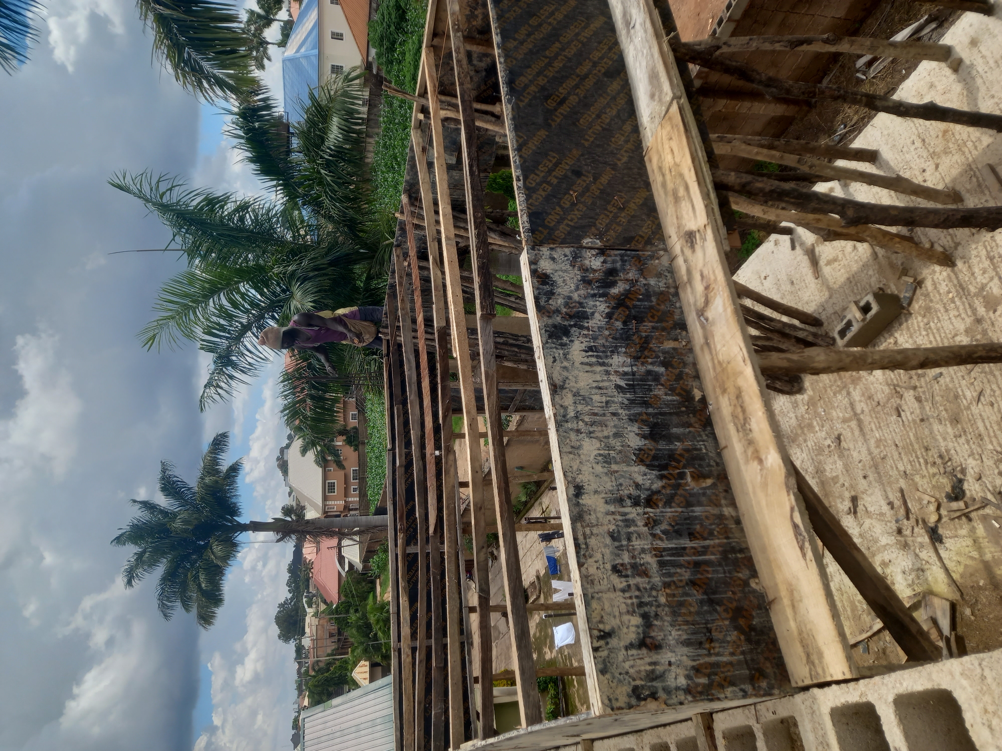 The image captures an outdoor construction scene in Gura Top, Nigeria, on a partly cloudy day. In the foreground, a partially constructed building features wooden scaffolding and framing, with plywood sheeting forming walls. A person wearing a hat and a purple shirt with a bag slung over their shoulder is visible working on the upper level of the structure. Laundry, including white and blue items, hangs to dry on a clothesline in the background, suggesting a residential area adjacent to the construction site. Palm trees and other lush vegetation surround the buildings, indicating a tropical or subtropical climate. Several houses with distinct roofing styles are visible in the distance, some with blue and red roofs. The overall scene depicts an active construction project within a developed neighborhood.