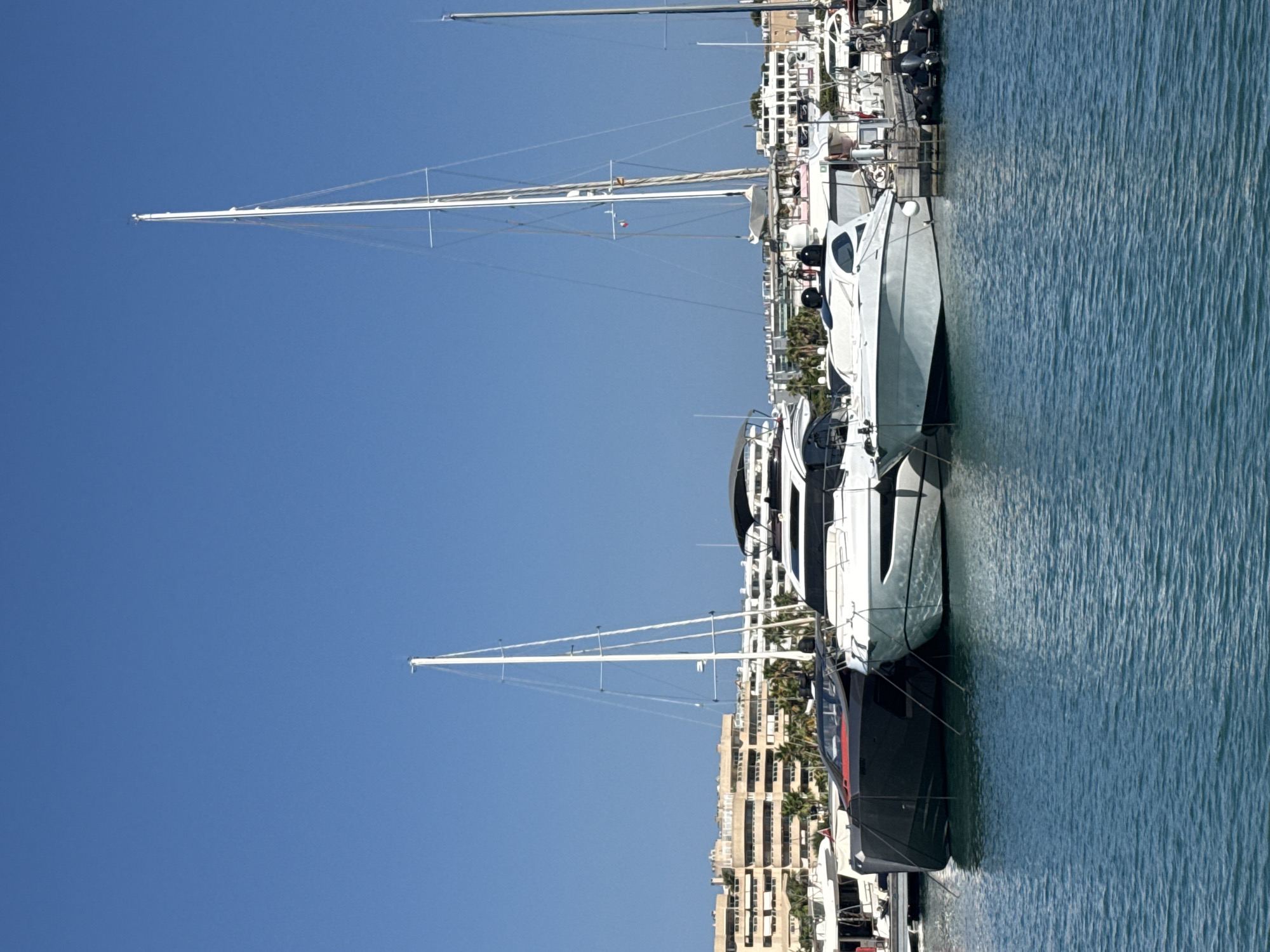 In Eivissa, Spain, a clear blue sky dominates the upper portion of the image. Several yachts are moored in a harbor. In the foreground, three motor yachts are visible. The leftmost yacht is dark gray with a red stripe. Next to it is a white yacht with black accents. To the right of the white yacht is another white motor yacht. Behind these yachts, a tall white sailboat mast extends upwards. In the background, a row of multi-story buildings with balconies lines the shore, interspersed with palm trees. The water in the foreground is a vibrant blue with small ripples.