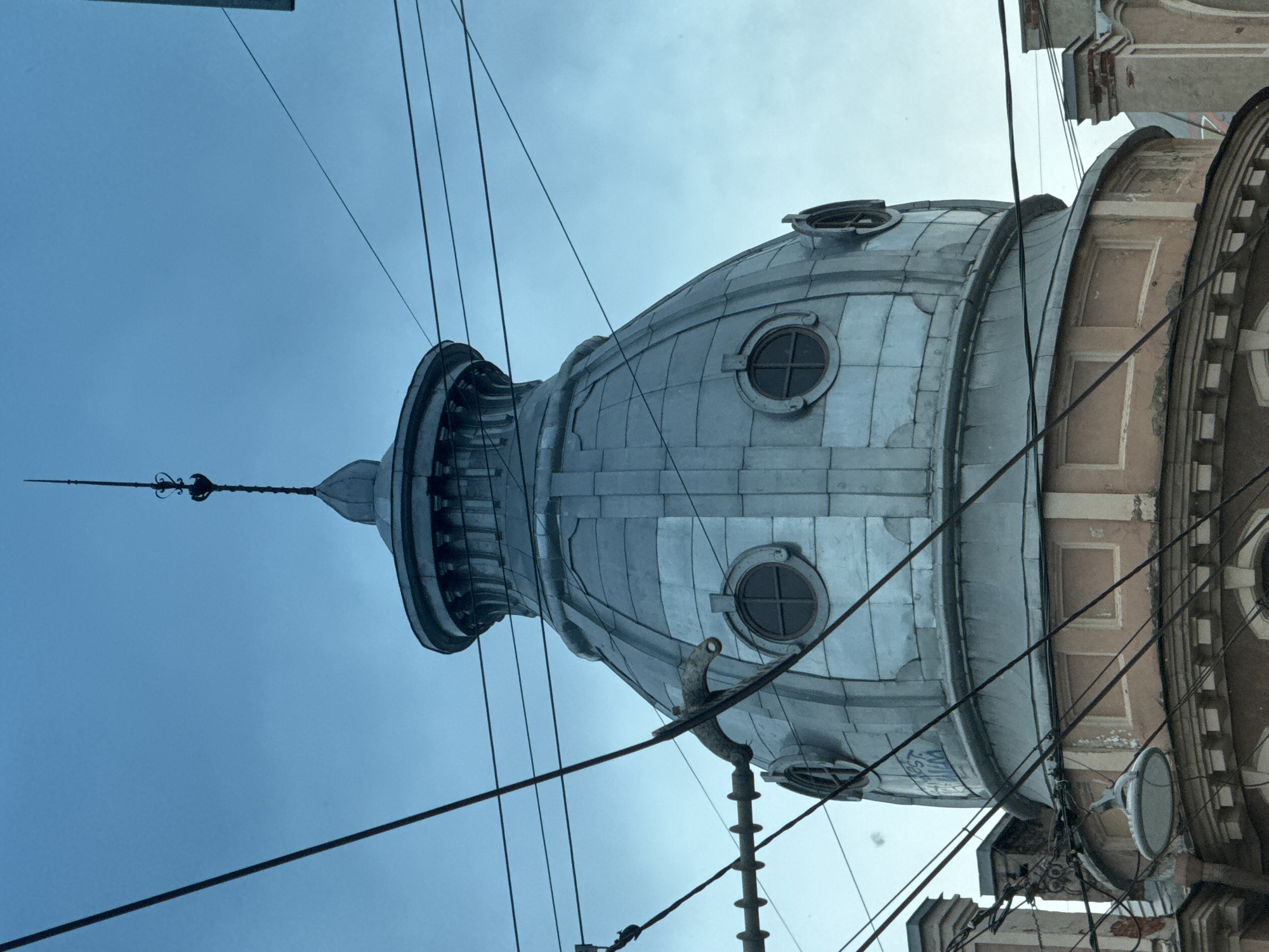 A photograph captures a close-up view of a domed building, likely in Saint Petersburg, Russia. The dome, constructed from metallic panels, has a weathered, silver-grey appearance. It is adorned with two circular windows on each side, featuring cross-shaped panes. A decorative spire tops the dome. The building's lower section, visible at the bottom, is constructed from a peach-colored plaster with some signs of wear. A street lamp hangs from a cluster of electrical wires that crisscross the frame, obscuring parts of the building. The sky above is a muted blue, suggesting overcast conditions or dusk. The overall impression is one of urban decay and industrial overhead infrastructure.