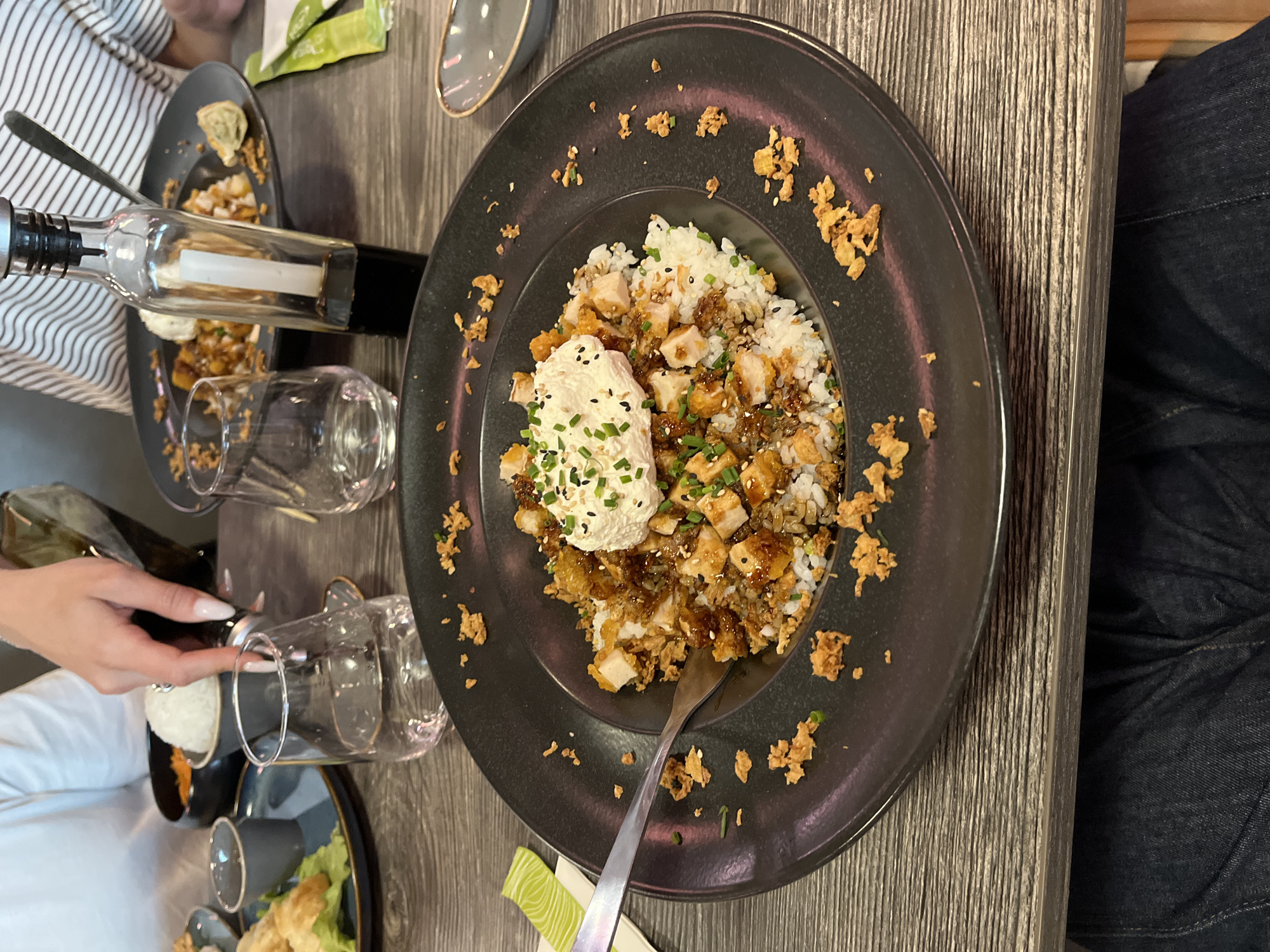 The image depicts a table setting, likely in a restaurant. A large, dark gray plate dominates the scene, featuring a rice-based dish topped with diced chicken, a dollop of creamy sauce adorned with black sesame seeds and chives, and scattered fried elements. A fork is positioned on the plate. Surrounding the main dish are several other elements, including glasses, a bottle with a pour spout, and another plate with remnants of food. A hand with manicured nails can be seen holding a glass. A person wearing jeans is also visible. The location is potentially Andrézieux-Bouthéon, France, based on the description prompt.