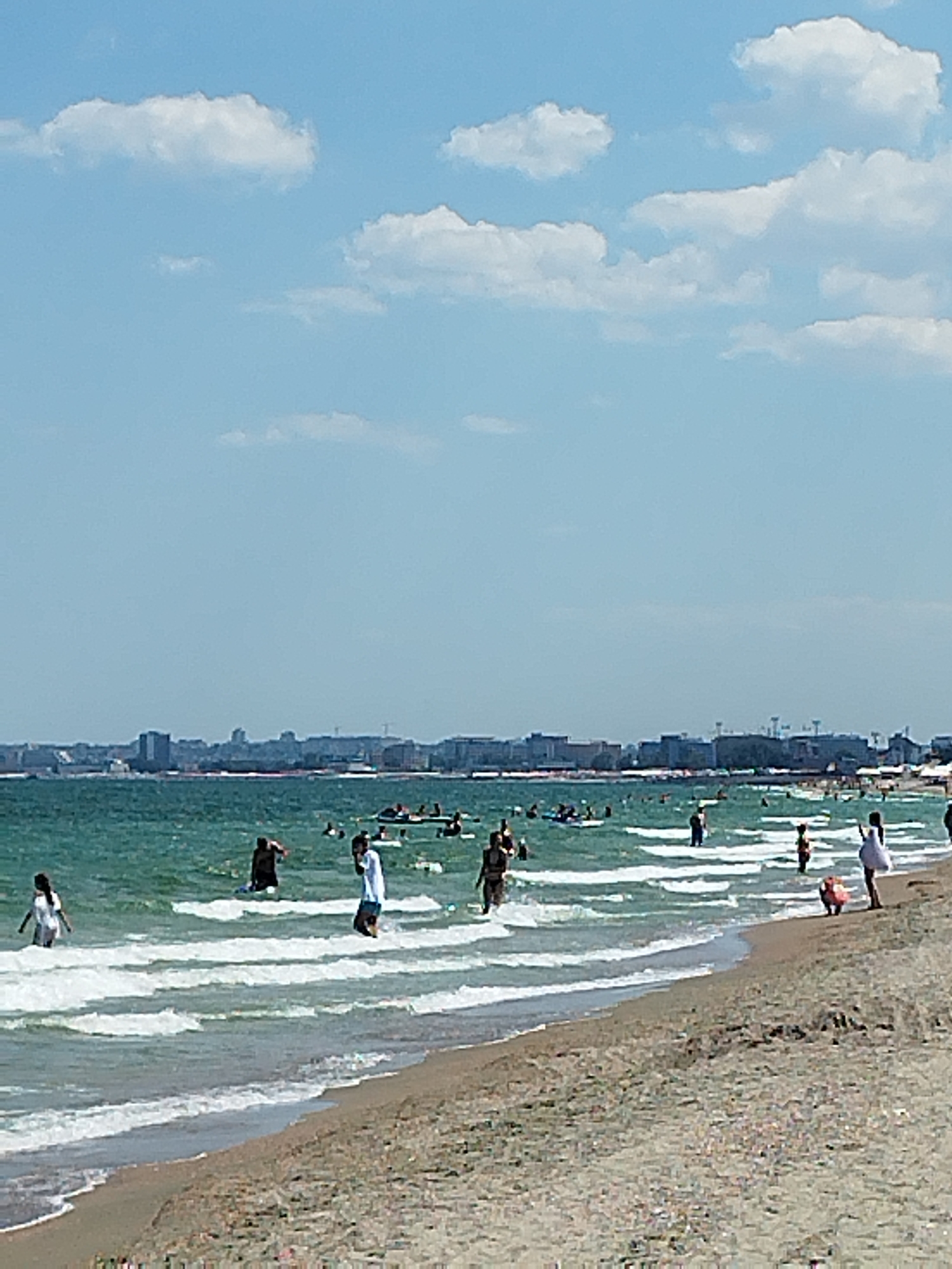 The image depicts a busy summer day at a beach in Constanța, Romania, likely situated along the Black Sea coast, with the city's skyline visible in the background. The water is a turquoise green, with white waves breaking on the shore. Numerous people are scattered throughout the water, engaging in activities such as swimming, wading, and playing. Some are visible on inflatable devices.
On the sandy beach, several individuals are present, including a woman in a white dress near the foreground, and a child in a red bathing suit. The beach itself appears to be well-populated, with more people visible further down the coastline. The sky is a clear blue with scattered white clouds. The city skyline in the distance features a mix of residential and commercial buildings, suggesting an urbanized coastal environment. The overall scene conveys a lively atmosphere typical of a popular seaside resort.