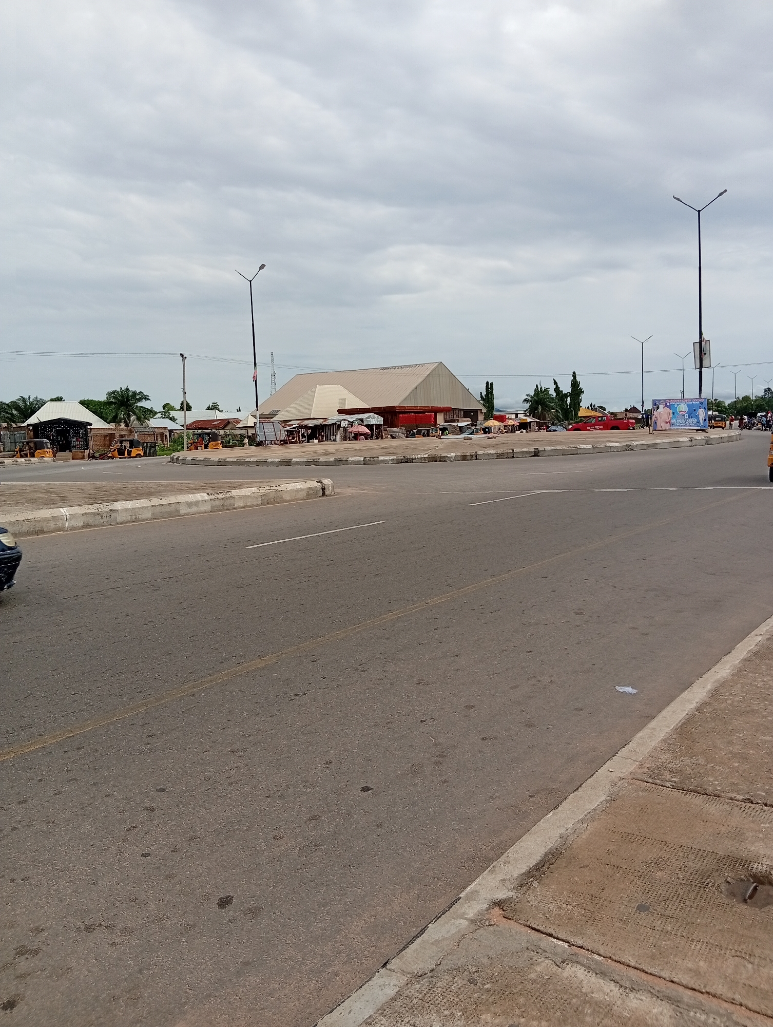 The image shows a wide, paved road with a concrete divider. On the other side of the divider are several buildings, some with awnings, and what appear to be market stalls. There are several yellow and orange "keke" (tricycle taxis) parked near the stalls, as well as a few other vehicles. In the background, there are more buildings and trees, and a large billboard with a picture of two men. Several lampposts are visible, and the sky is overcast, suggesting a cloudy day. The overall scene depicts a busy public area, likely a market or transportation hub, in Jalingo, Nigeria. The time of day appears to be daytime, but the cloudy weather obscures direct sunlight. The presence of the keke taxis and market stalls provides clear context for the location being a place of commerce and transportation.