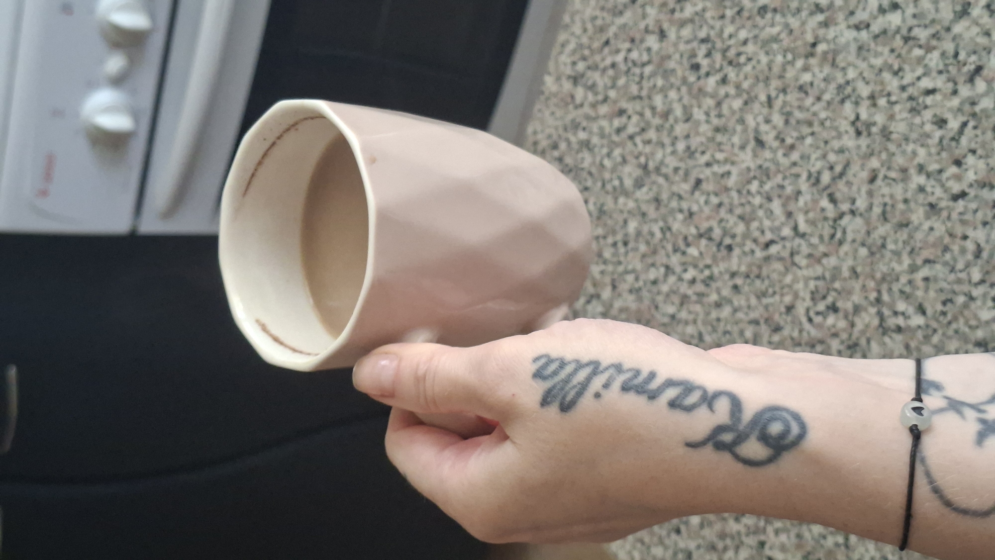 A person's left hand, tattooed with the name "Kamilla" and a floral design, holds a pink ceramic mug filled with a light brown beverage. The mug has a faceted exterior and a rim with a subtle darker band. A thin black string bracelet with a white bead featuring a black heart charm is visible on the person's wrist. The background shows a dark surface on the left, possibly a cabinet or appliance, and a white oven with knobs in the upper right corner. The surface beneath the hand and mug is a speckled countertop. The location is not Myadzyel, Belarus.