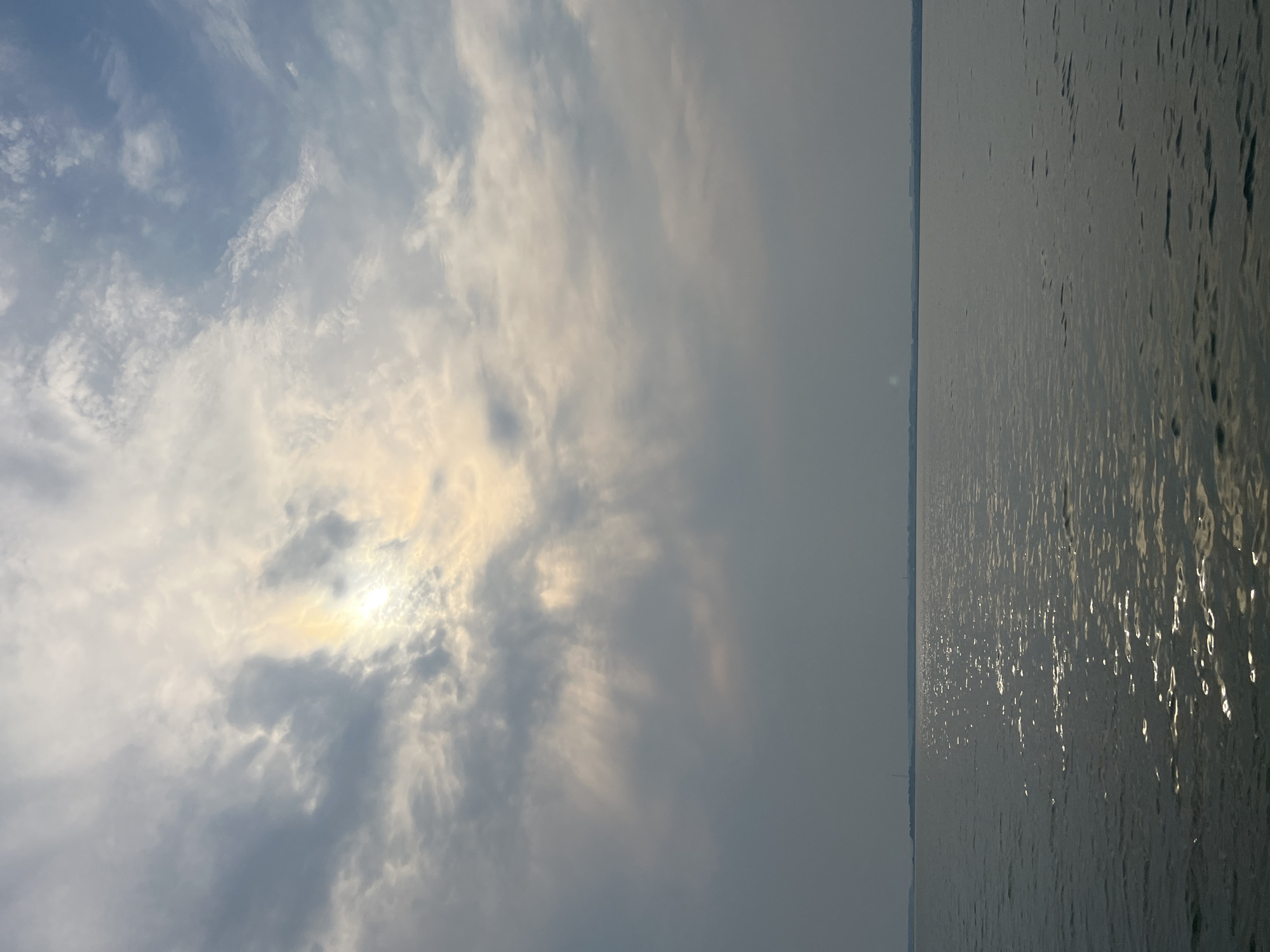 The image depicts a wide expanse of calm water, likely a lake or a large river, stretching towards a distant, hazy horizon. The water's surface is disturbed by gentle ripples, reflecting the light in shimmering patches. A faint shoreline with sparse vegetation is visible in the distance, under a diffused, overcast sky. The sky itself is filled with a mix of clouds, ranging from dark, heavy masses to lighter, wispy formations. The sun is partially obscured by clouds, casting a soft, diffused light and creating a subtle halo effect. There are no people, animals, or man-made structures clearly visible in the immediate foreground or the water. The overall impression is one of a serene, natural landscape, with a subdued, atmospheric quality. The location in Novopichugovo, Russia, is a detail provided externally and not discernible from the image itself.