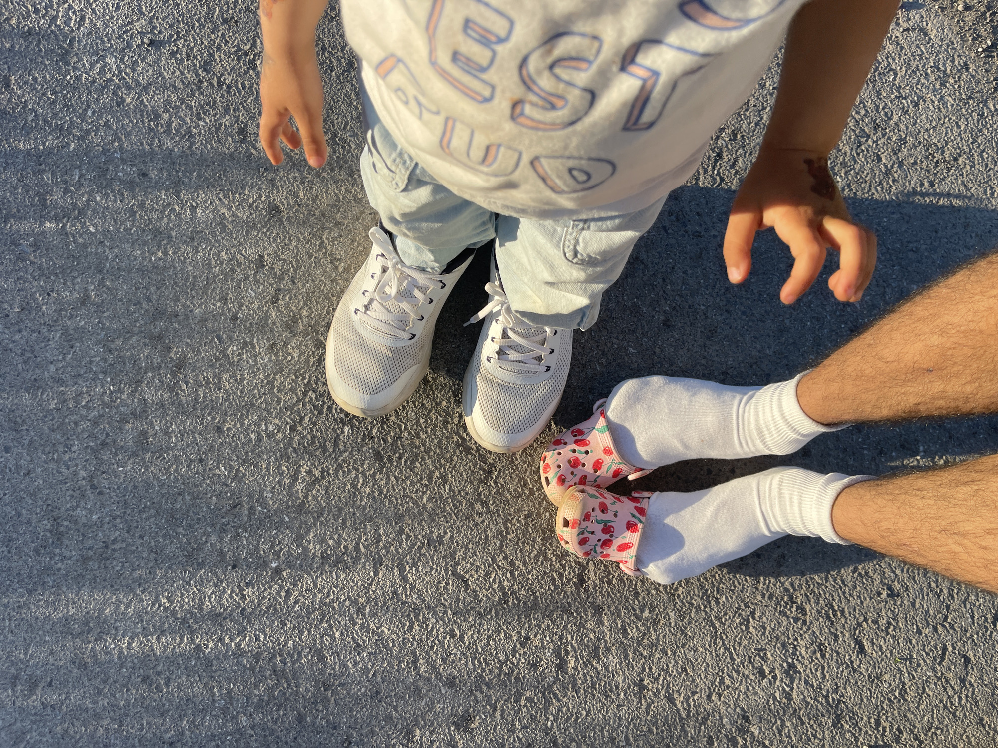 A top-down shot shows two people standing on a concrete surface. One person, presumably a child, is wearing white sneakers, light blue pants, and a white t-shirt. The other person, likely an adult, is wearing white socks and pink clogs with a strawberry pattern. The location is unknown.