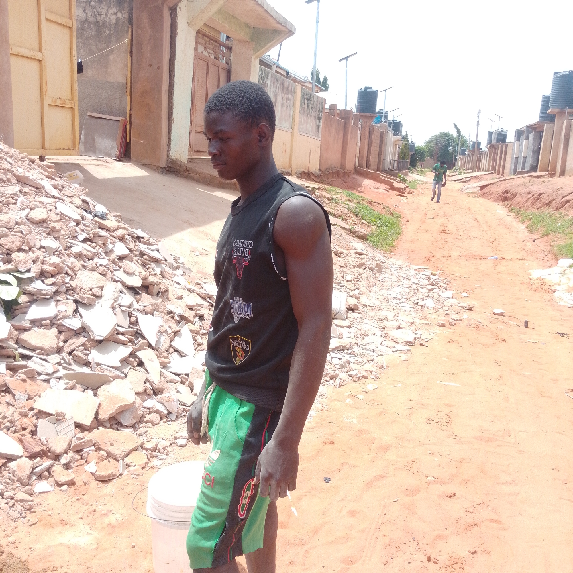 In Tumfure, Nigeria, a young Black male stands on a dirt road next to a pile of construction debris. He is wearing a black sleeveless shirt with graphics and green shorts. A white bucket is visible on his left. The background shows buildings and water tanks, suggesting a residential area. The sky is bright and overcast.