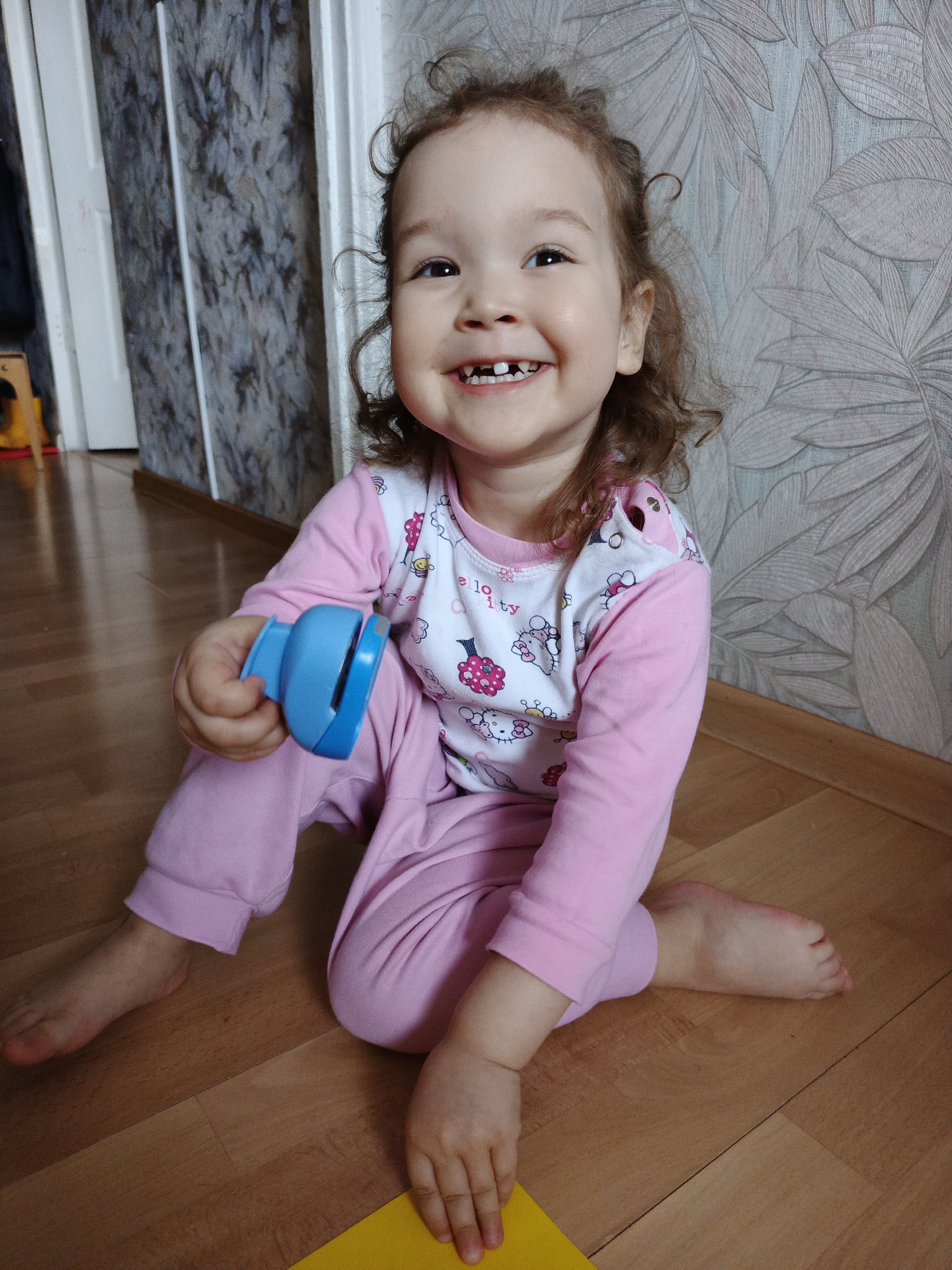 A young child is sitting on a wooden floor, indoors. The child is wearing pink pajamas with a Hello Kitty design on the shirt. They are holding a blue, plastic object in their left hand and a yellow piece of paper is visible under their right hand. The child is smiling, revealing some missing teeth. The walls behind the child are decorated with patterned wallpaper in shades of gray and white. A doorway is partially visible to the left of the child.