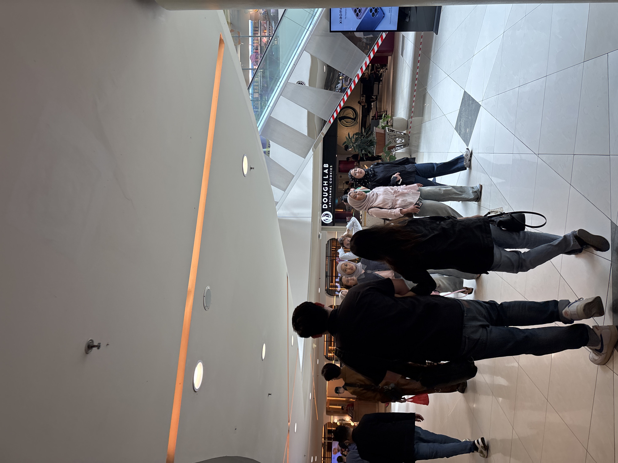 This image depicts the interior of a modern shopping mall, likely located in West Jakarta, Indonesia. A group of people are walking on a polished tiled floor. In the foreground, a man and a woman, viewed from behind, are walking away from the camera. The man is wearing a black t-shirt and jeans, while the woman is in a black long-sleeved shirt and jeans. Ahead of them, several other individuals are visible, including women wearing headscarves.
To the right, an escalator with glass railings ascends towards an upper level, where a supermarket or retail store with shelves stocked with products can be seen. A large advertisement featuring a smartphone is displayed on a vertical screen to the right of the escalator.
In the background, a store sign reads "DOUGH LAB ARTISANAL COOKIES" above an entrance. The ceiling of the mall features recessed lighting and accent lighting strips. The overall environment suggests a busy commercial space.