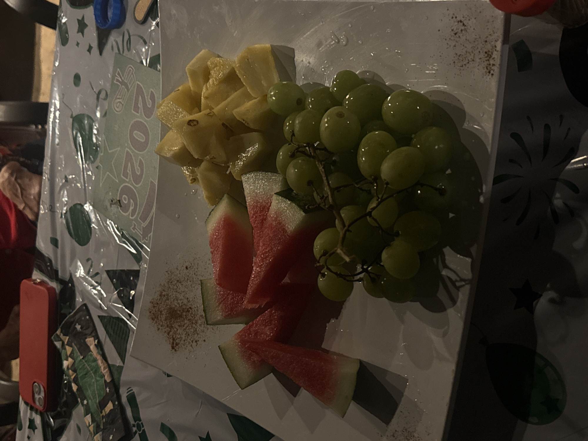 The image shows a plate of fruit containing pineapple, grapes, and watermelon. The plate rests on a festive tablecloth decorated with images of stars, balloons, and the year "2026," along with a graduation cap. To the left of the plate, a red phone is visible. The location is Southlake, United States of America.