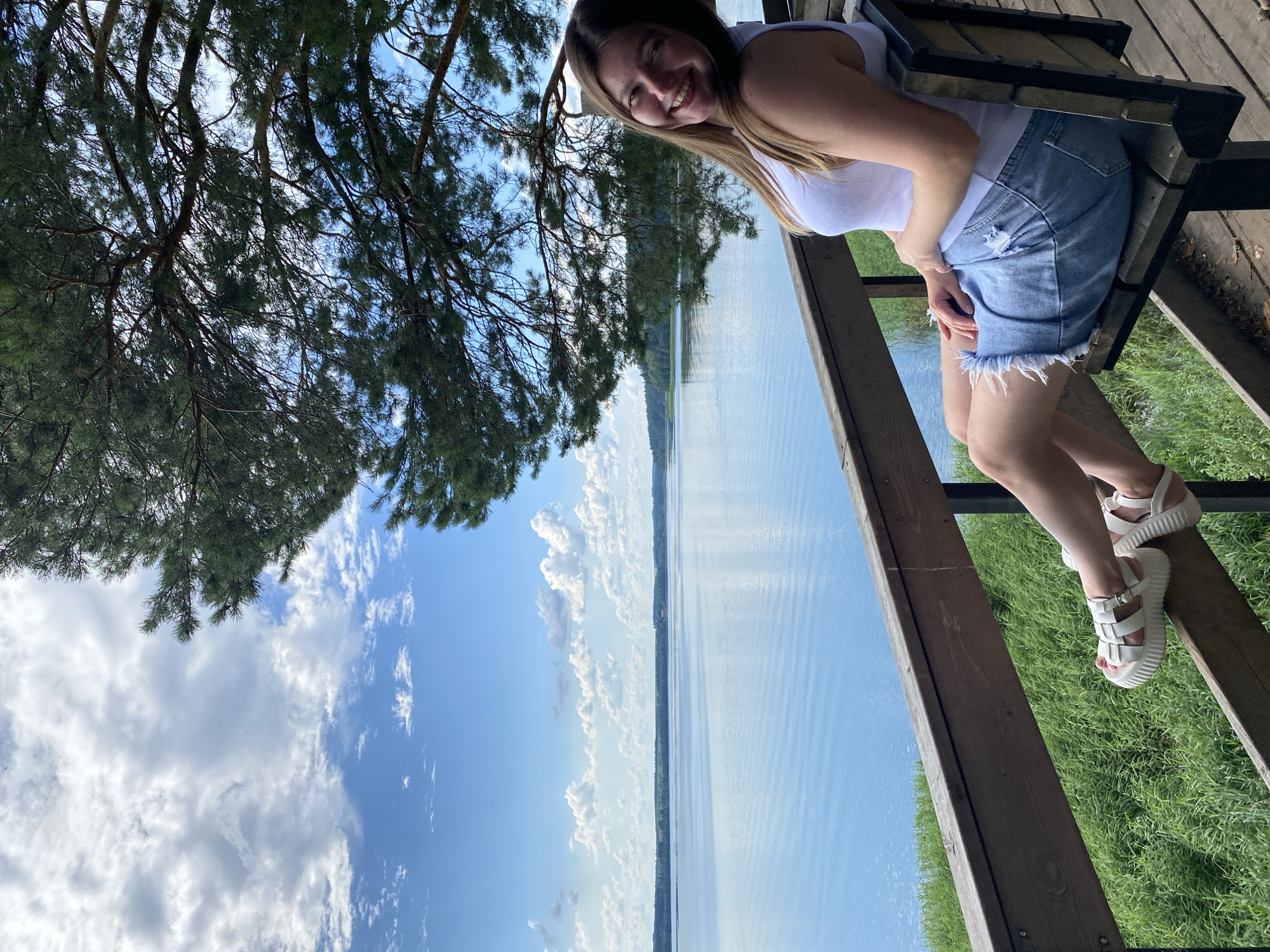 A young woman with long brown hair smiles at the camera while sitting on a wooden bench overlooking a large body of water. She is wearing a white tank top and a denim skirt with a frayed hem. White platform sandals are on her feet, which are resting on the railing of the bench. The bench is positioned on a wooden platform, with dense green grass growing on the bank below. Tall trees with green needles extend into the frame from the right, their branches creating a natural canopy. The water is calm and reflects the blue sky, which is dotted with white clouds. The shoreline in the distance is lined with trees. The scene appears to be a scenic overlook, possibly in Votkinsk, Russia, given the surrounding natural landscape.